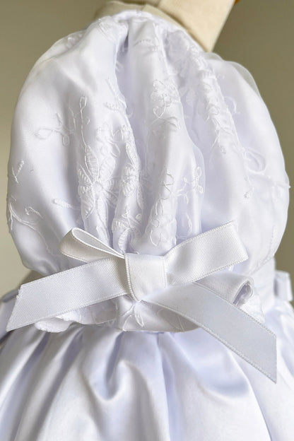 Detailed view highlights white satin flower girl dress with lace details and a large bow. Also suitable as a communion dress.