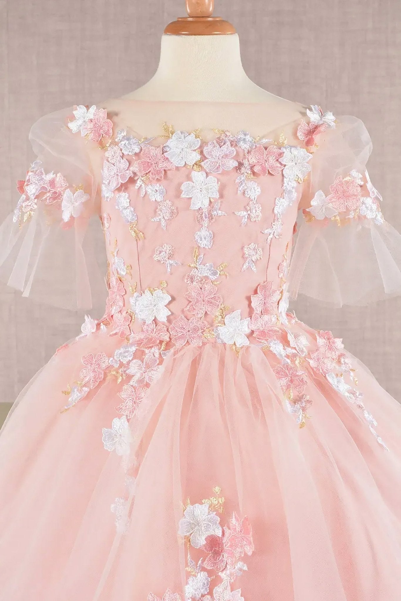 Front zoom view of a blush kid ballgown with 3D floral applique, puff sleeves, and long back hem