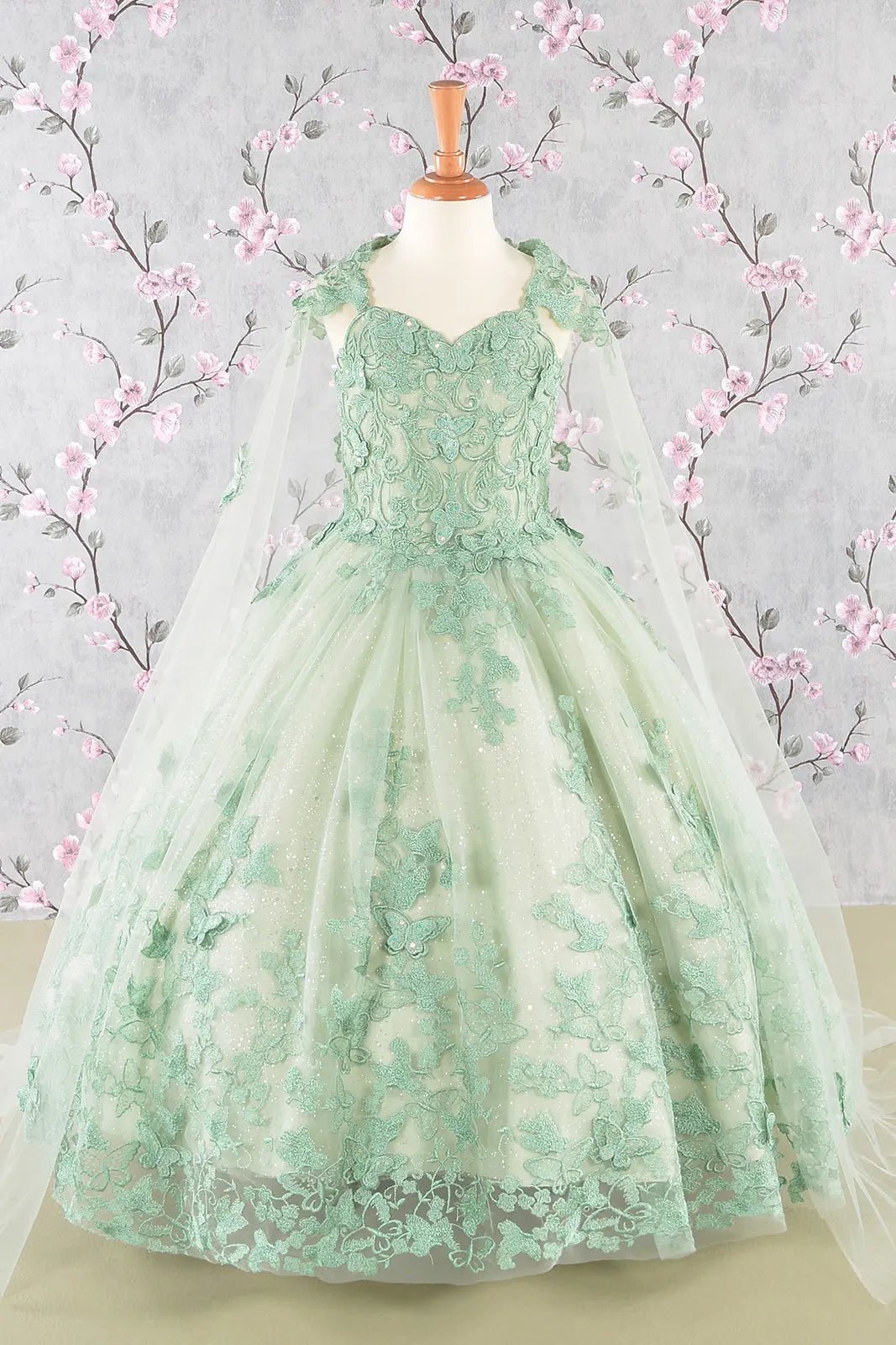 Sage green kids mini quinceañera mini quince dress with 3D butterfly appliques, sequin-embellished bodice, glitter tulle skirt, and flowing mesh cape.