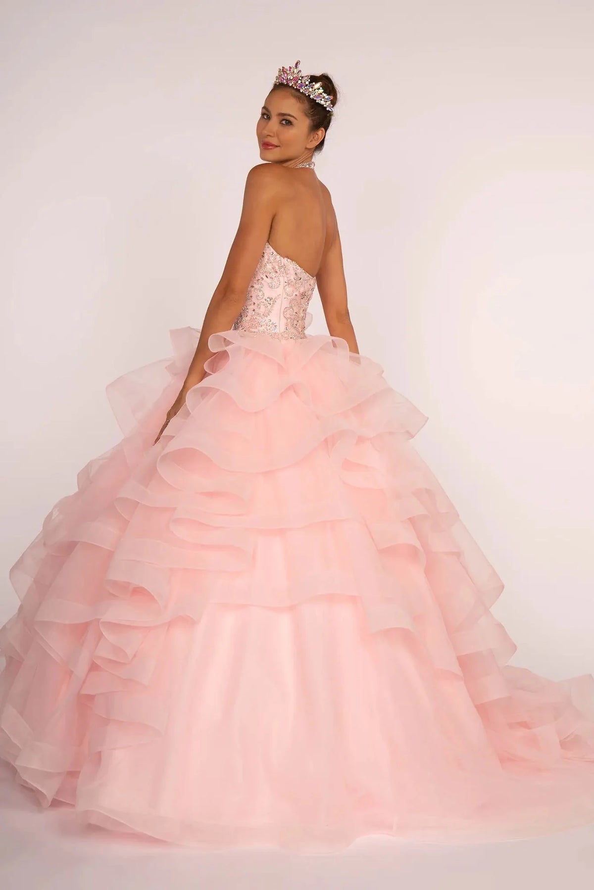 From a full back view, this Baby Pink quinceañera dress displays a low beaded back above wide horizontal ruffles.