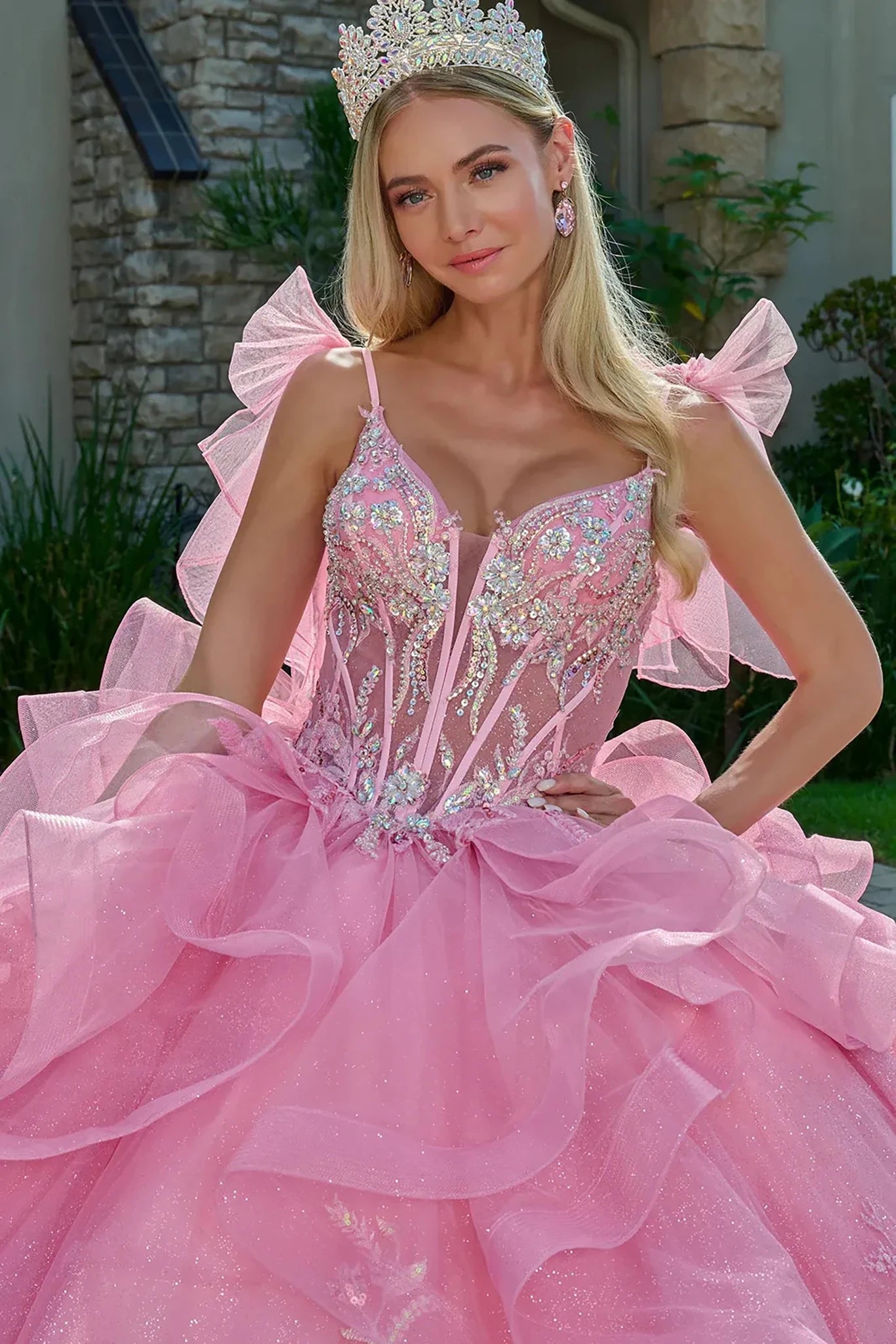 Woman in a pink gown with intricate detailing and a tiara, standing outdoors.