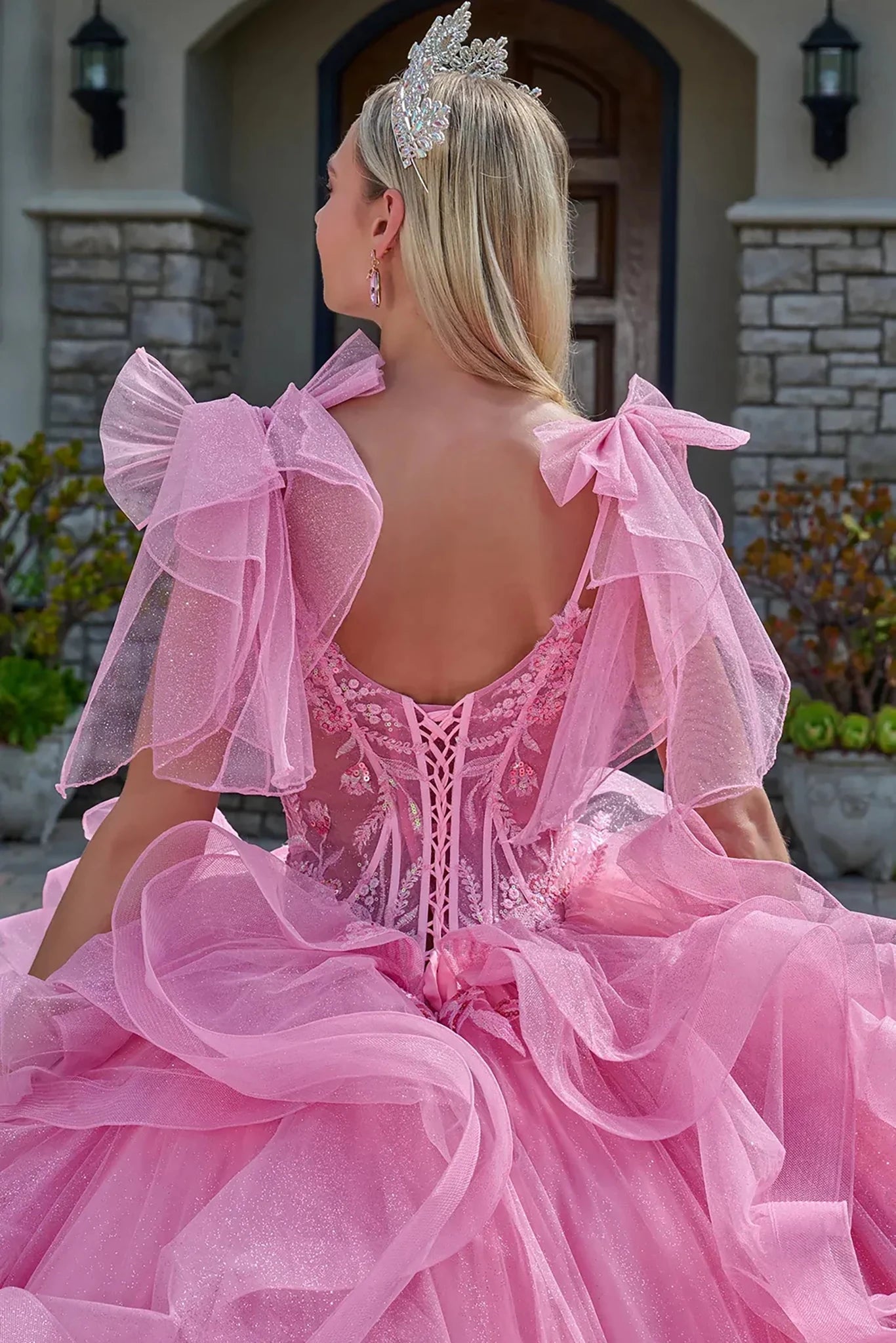 Woman in a pink dress with ruffled sleeves and a tiara, standing in front of a stone building.