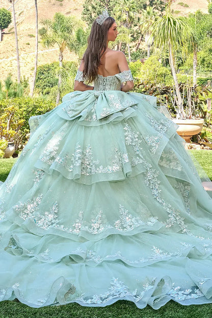Woman in a sage green ball gown with floral details and long ruffle train standing outdoors.