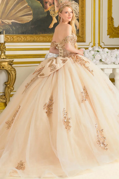 Woman in a beige and gold ball gown standing in an ornate room with gold frames and white flowers.