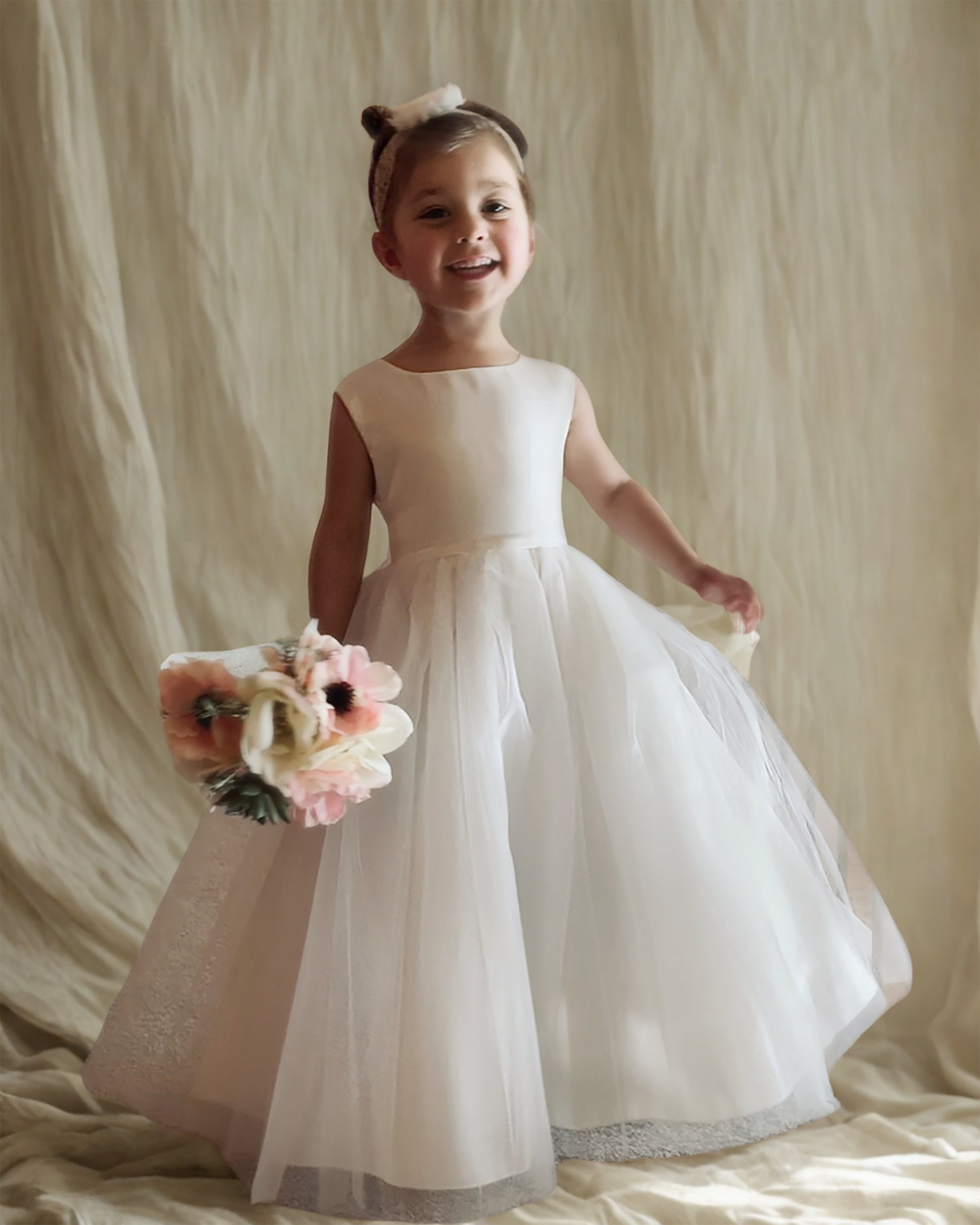 Young girl in a white dress holding flowers against a beige curtain background