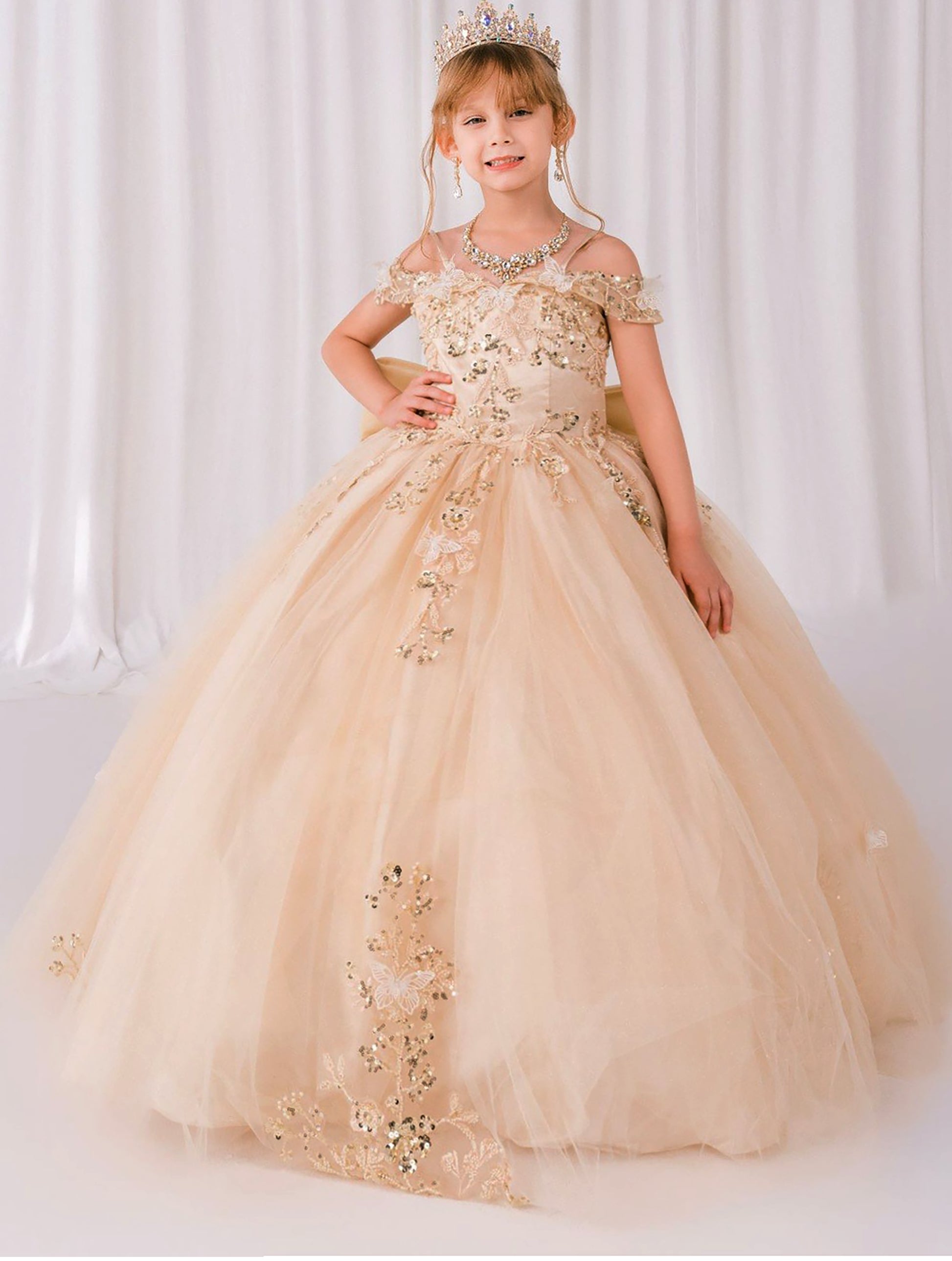 Young girl in a gold dress with floral embellishments against a white curtain background