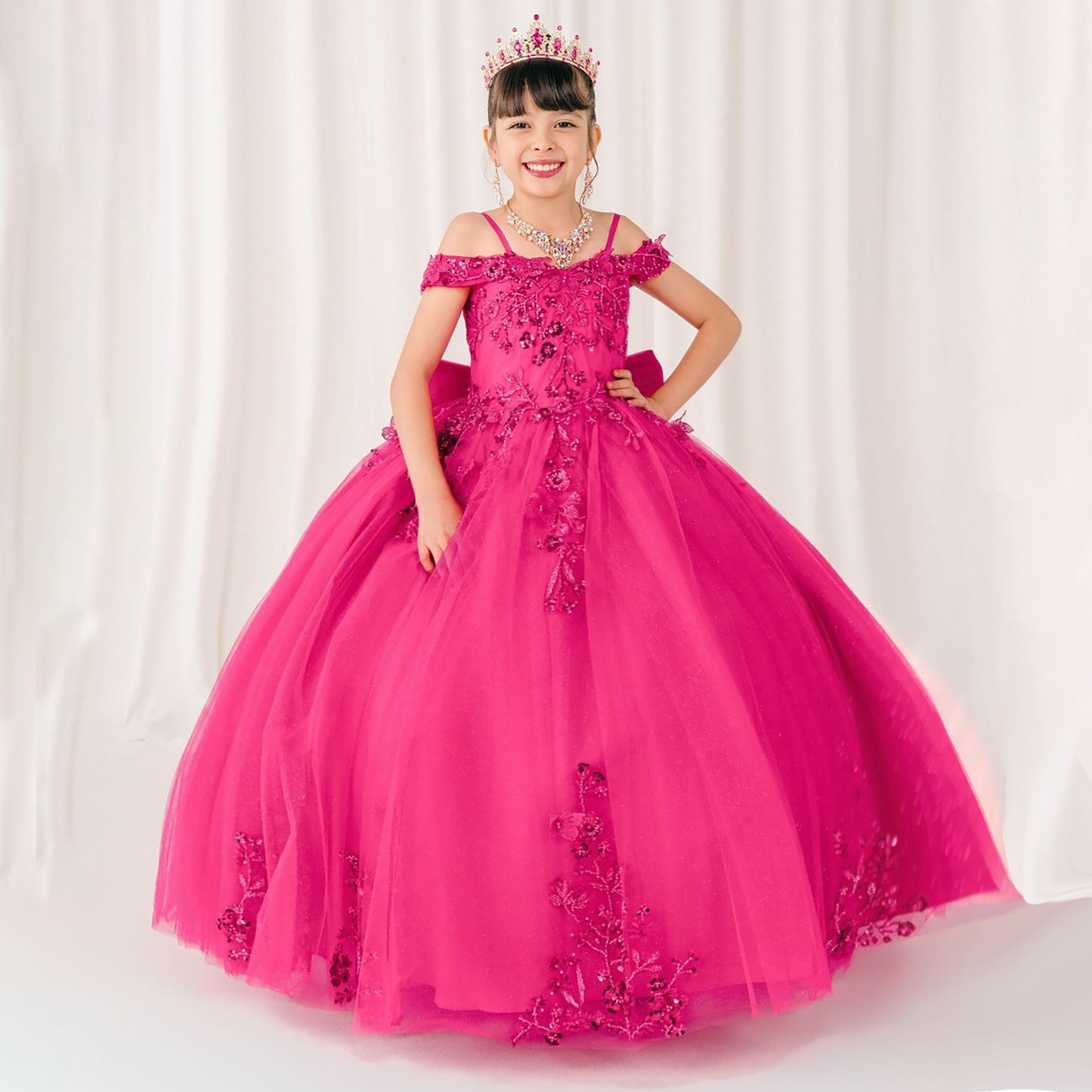 Young girl in a fuchsia dress with floral details and a tiara against a white curtain background