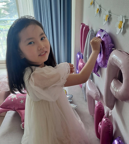 Young girl wearing a tiara in a white dress standing in a room with decorative balloons and streamers.