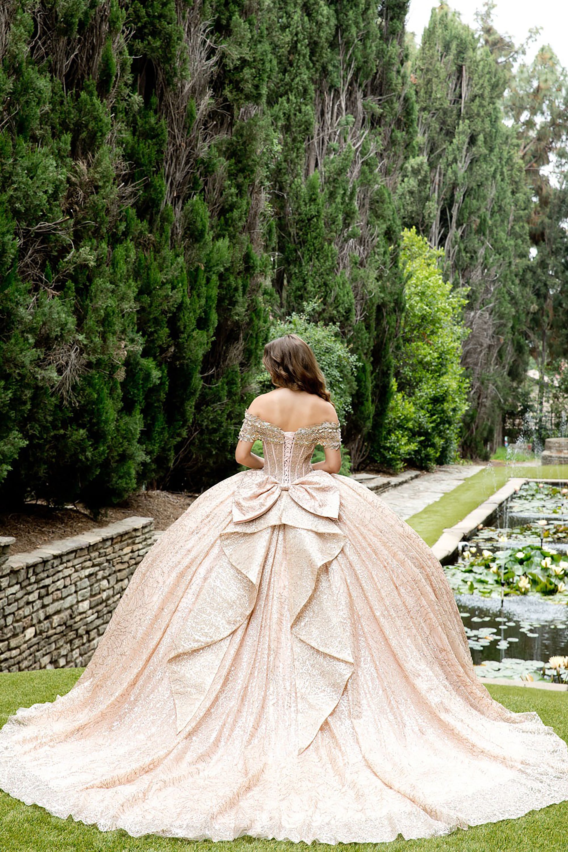 Woman in a rose gold dress standing in a garden with trees and a pond.