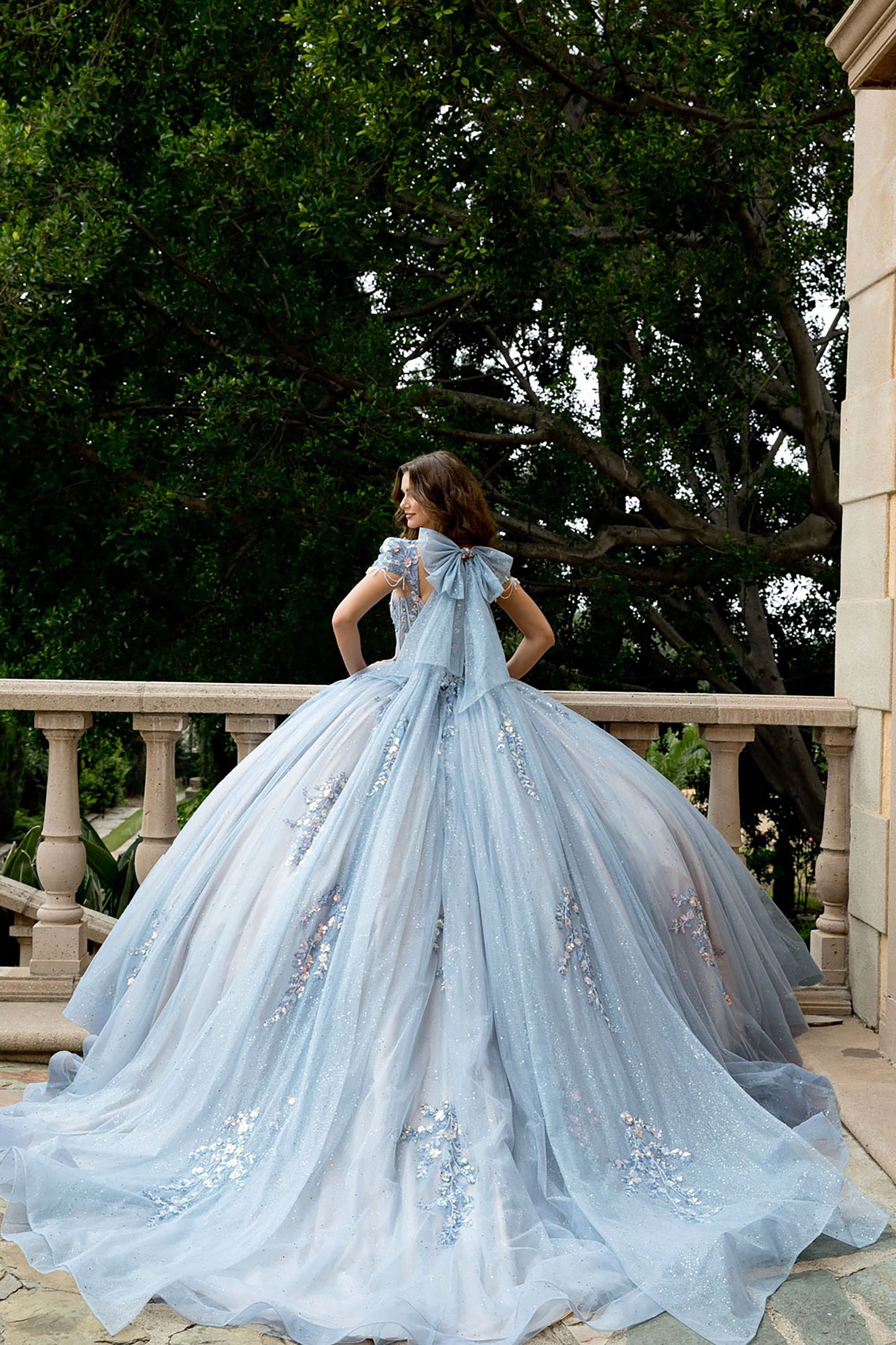 Woman in a light blue gown standing on a balcony with trees in the background