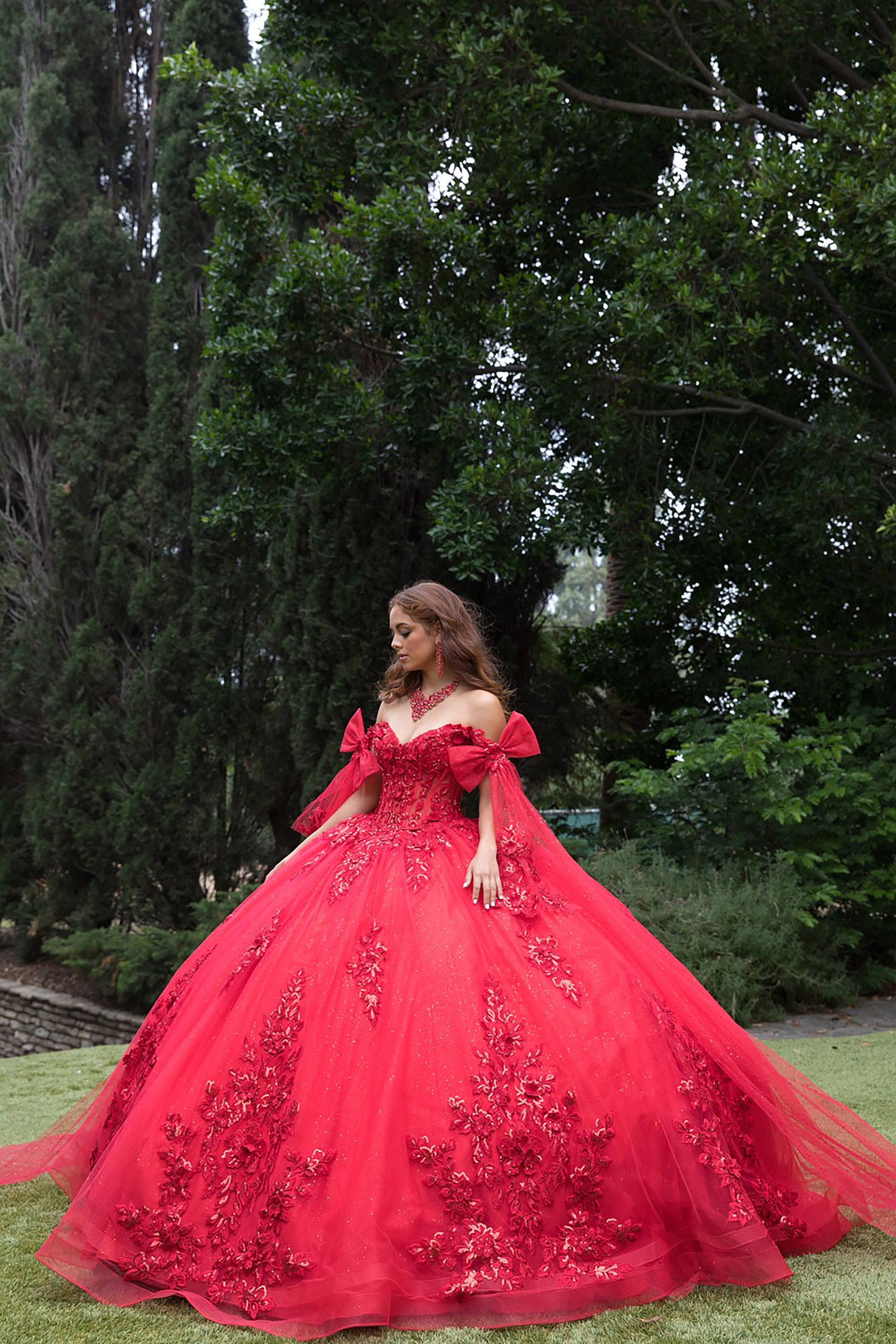 Woman in a red ball gown with floral embroidery standing outdoors among trees.