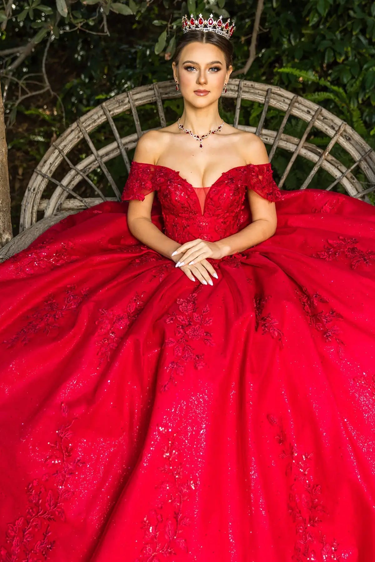 Woman in a red off-shoulder gown with a tiara, sitting outdoors.