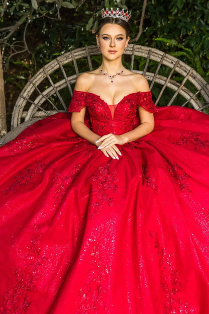 Woman in a red off-shoulder gown with a tiara, sitting outdoors.