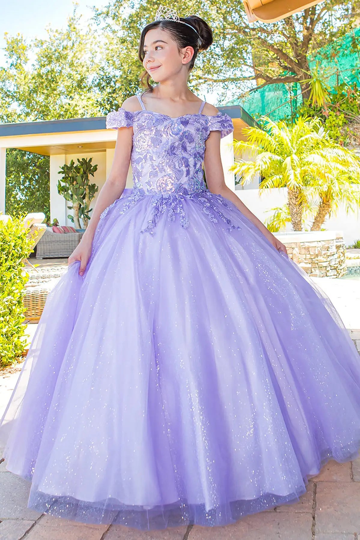 Young girl in a lilac ball gown with 3D appliques and off shoulder  standing outdoors with trees and a pool in the background.