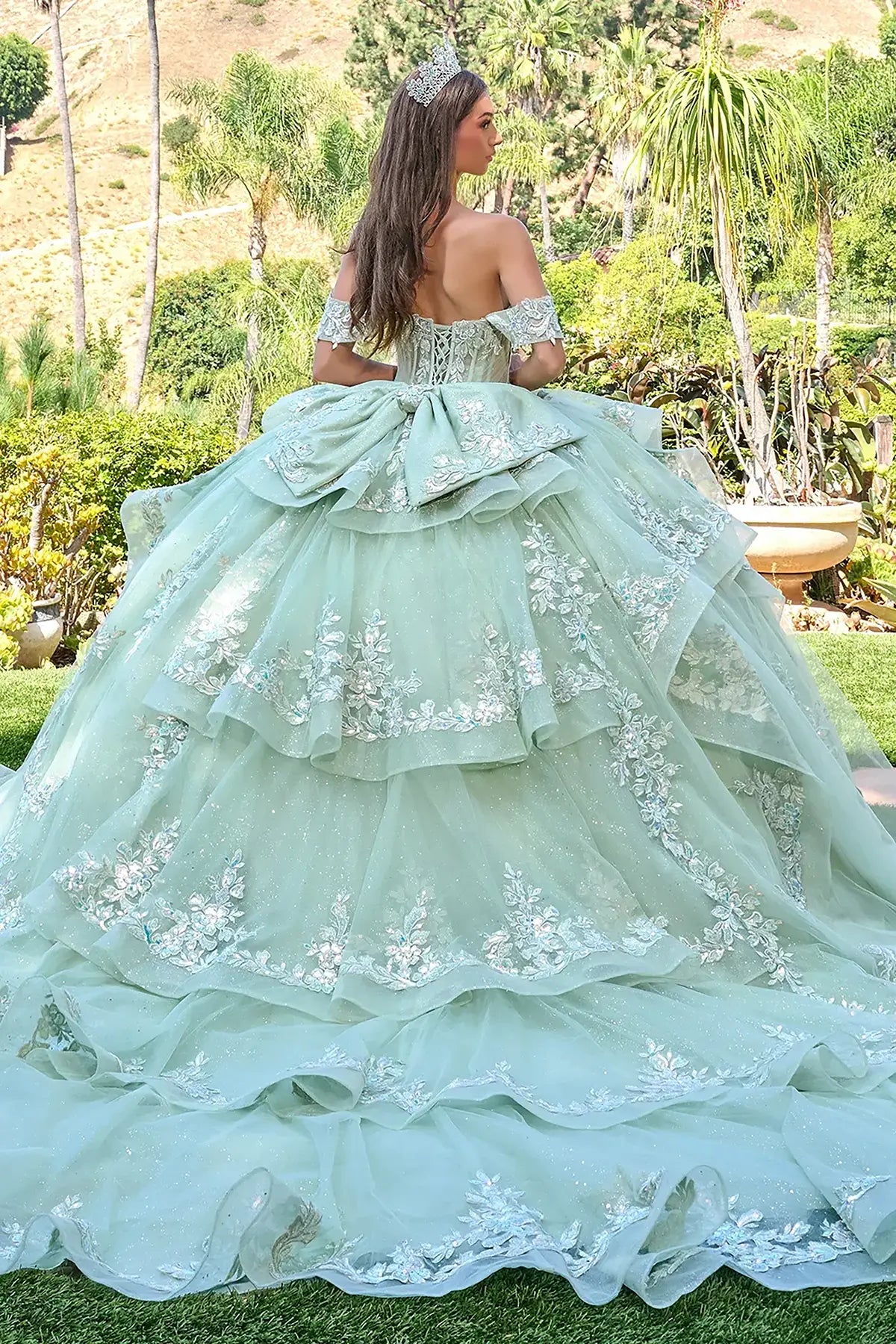 Woman in a sage green ball gown with floral details and long ruffle train standing outdoors.