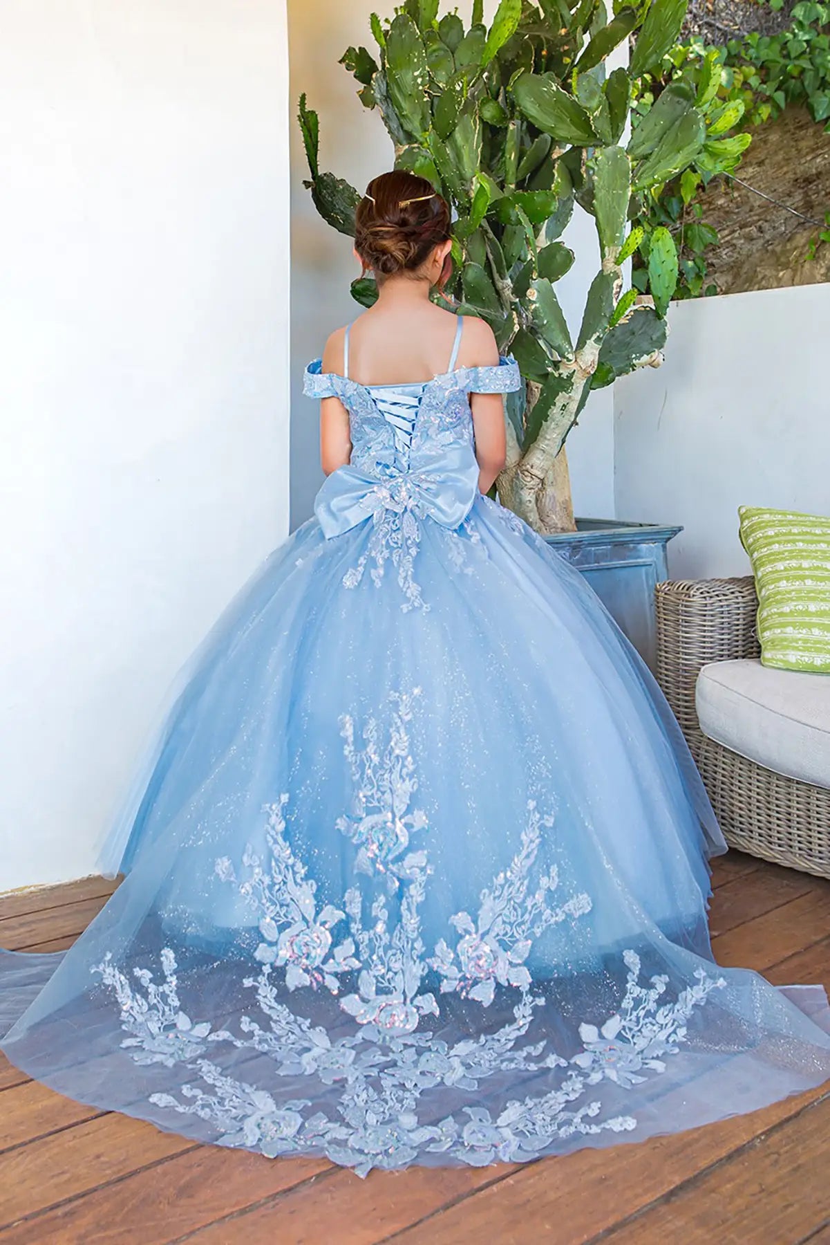 Kid in a blue off-shoulder ball gown dress with floral details standing next to a potted plant. the dress has long train