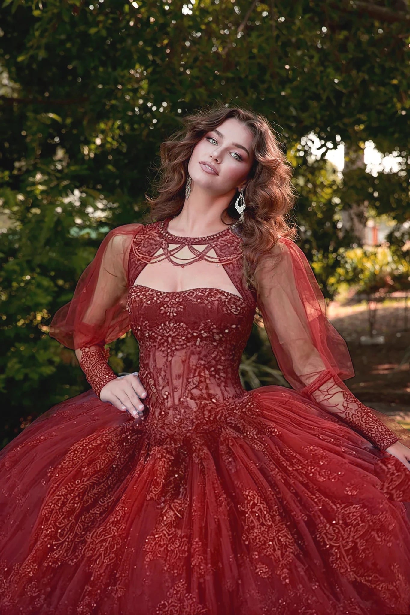 Woman in a red, intricately designed dress with see though long sleeves standing outdoors with greenery in the background