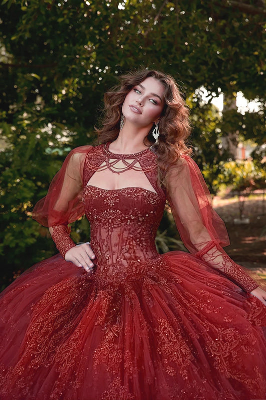 Woman in a red, intricately designed dress with see though long sleeves standing outdoors with greenery in the background