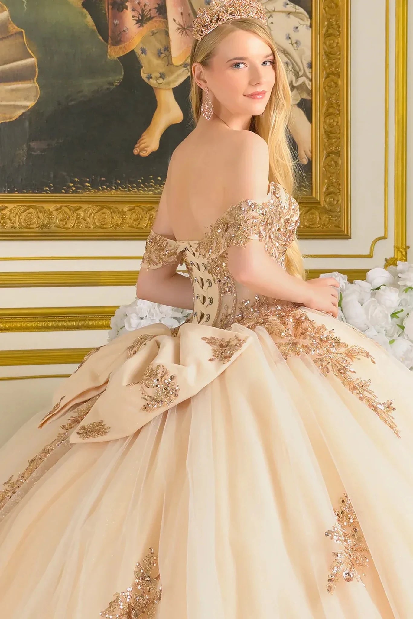 Woman in a beige and gold formal gown with intricate detailing, standing in an ornate room.