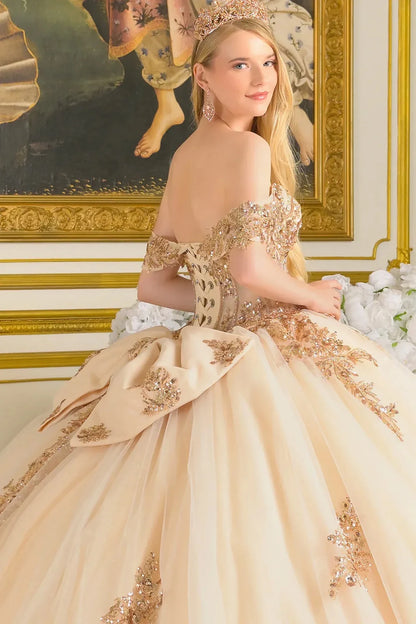Woman in a beige and gold formal gown with intricate detailing, standing in an ornate room.