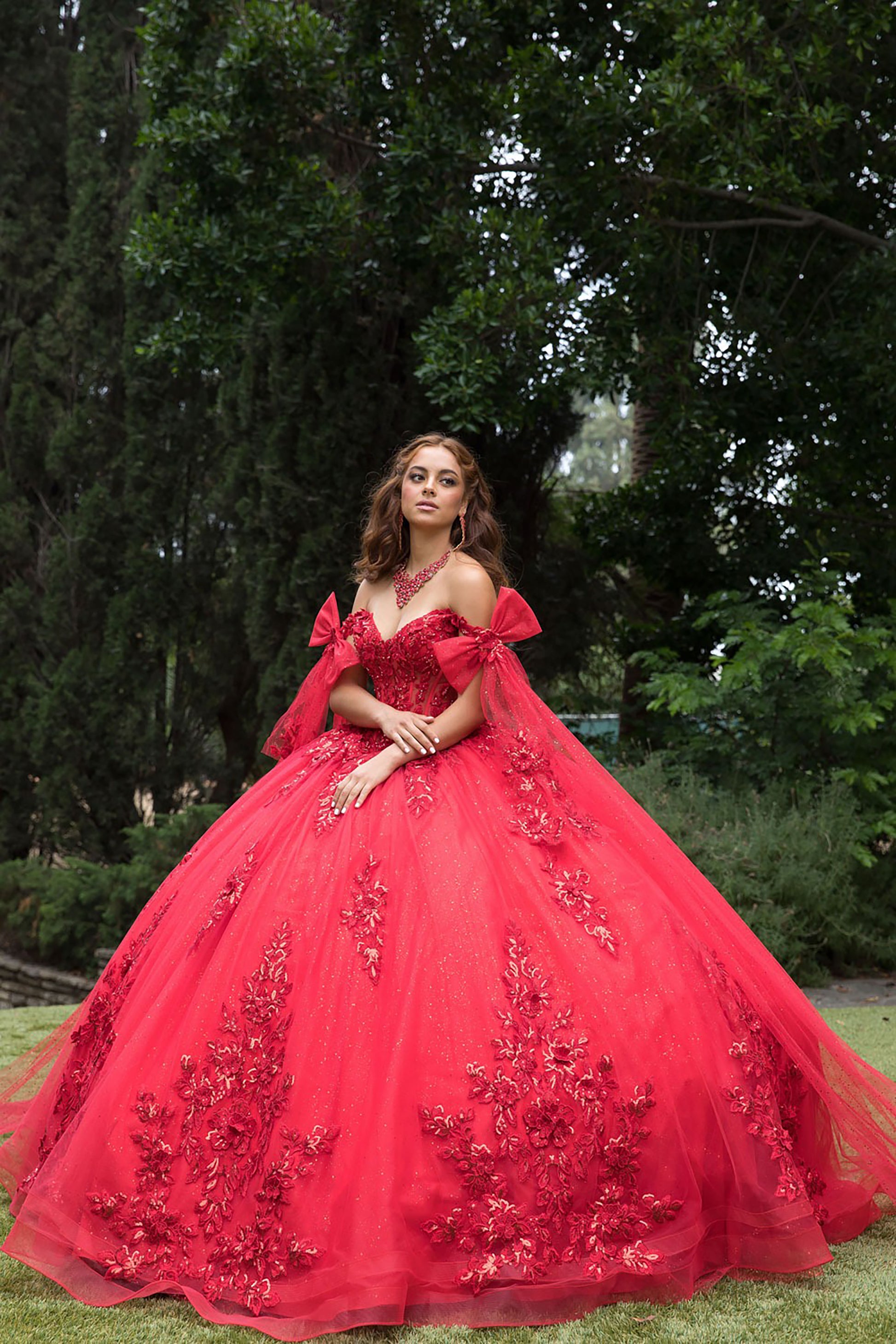 Woman in a red ball gown with floral details standing outdoors among trees.