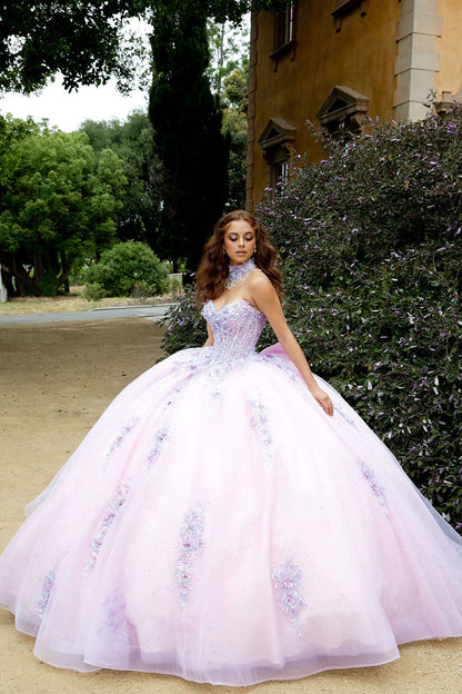 Woman in a pink ball gown with floral patterns standing outdoors near a building and trees.