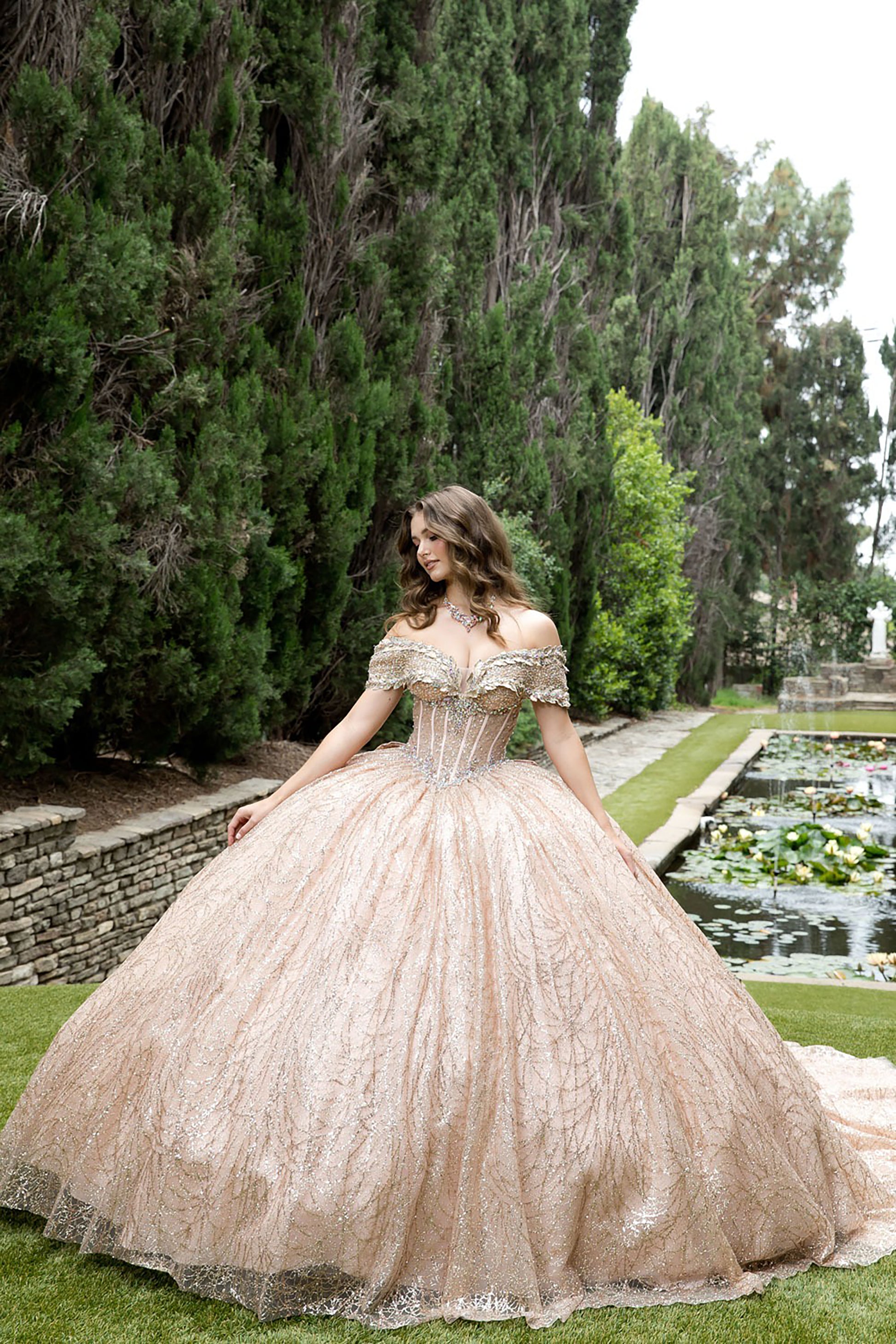 Woman in a rose gold lace dress standing outdoors with trees and water features in the background
