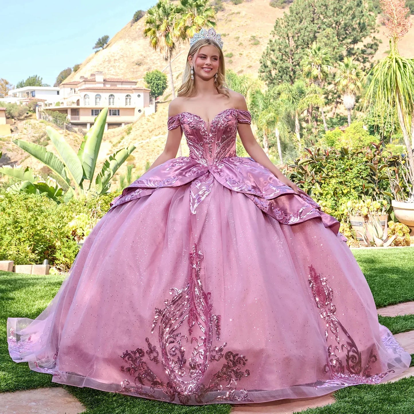 Woman in a dusty rose quinceañera dress standing outdoors with greenery and hills in the background