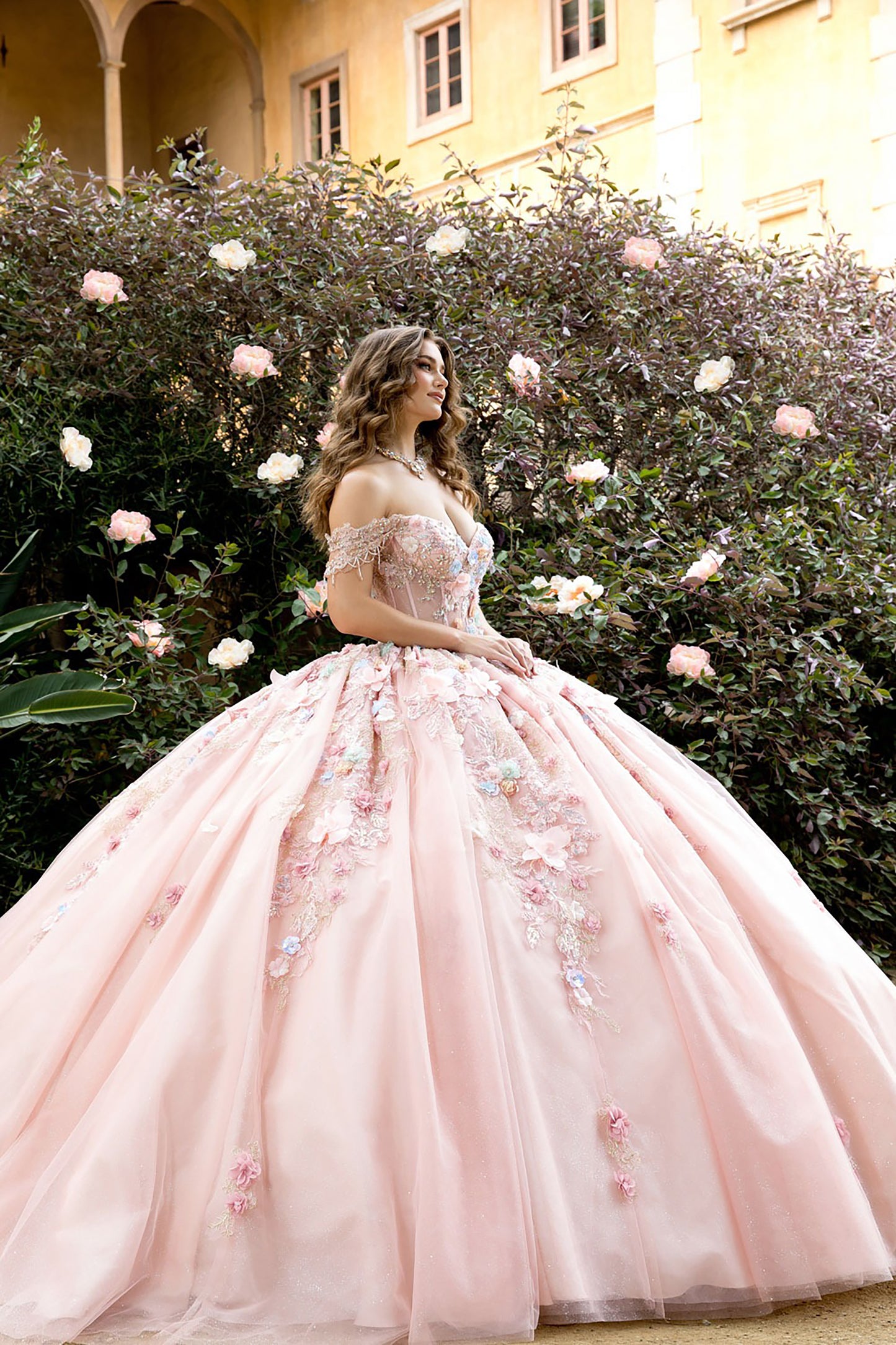 Woman in a pink floral dress standing in front of a flowering bush with a building in the background