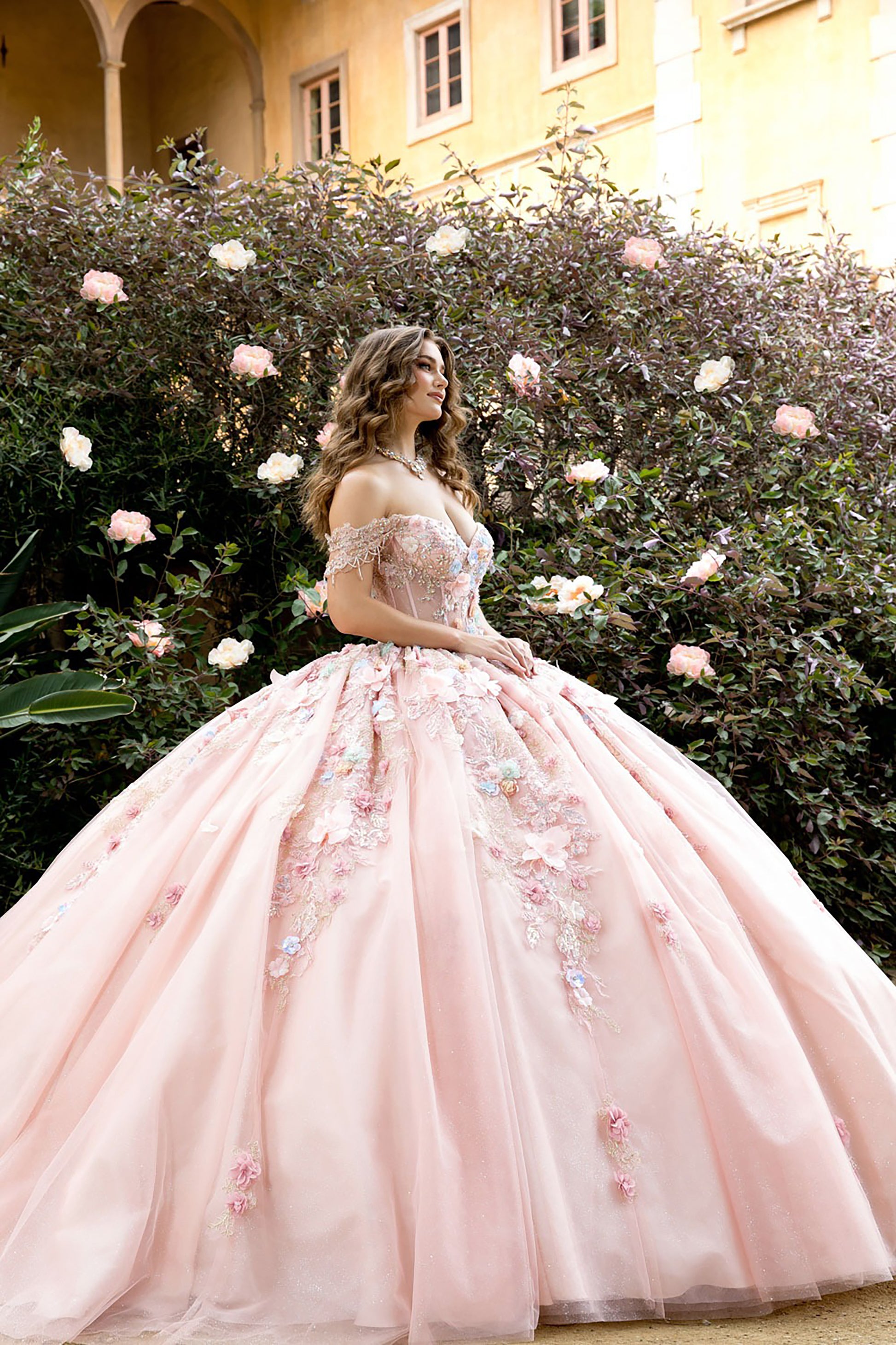 Woman in a pink floral dress standing in front of a flowering bush with a building in the background