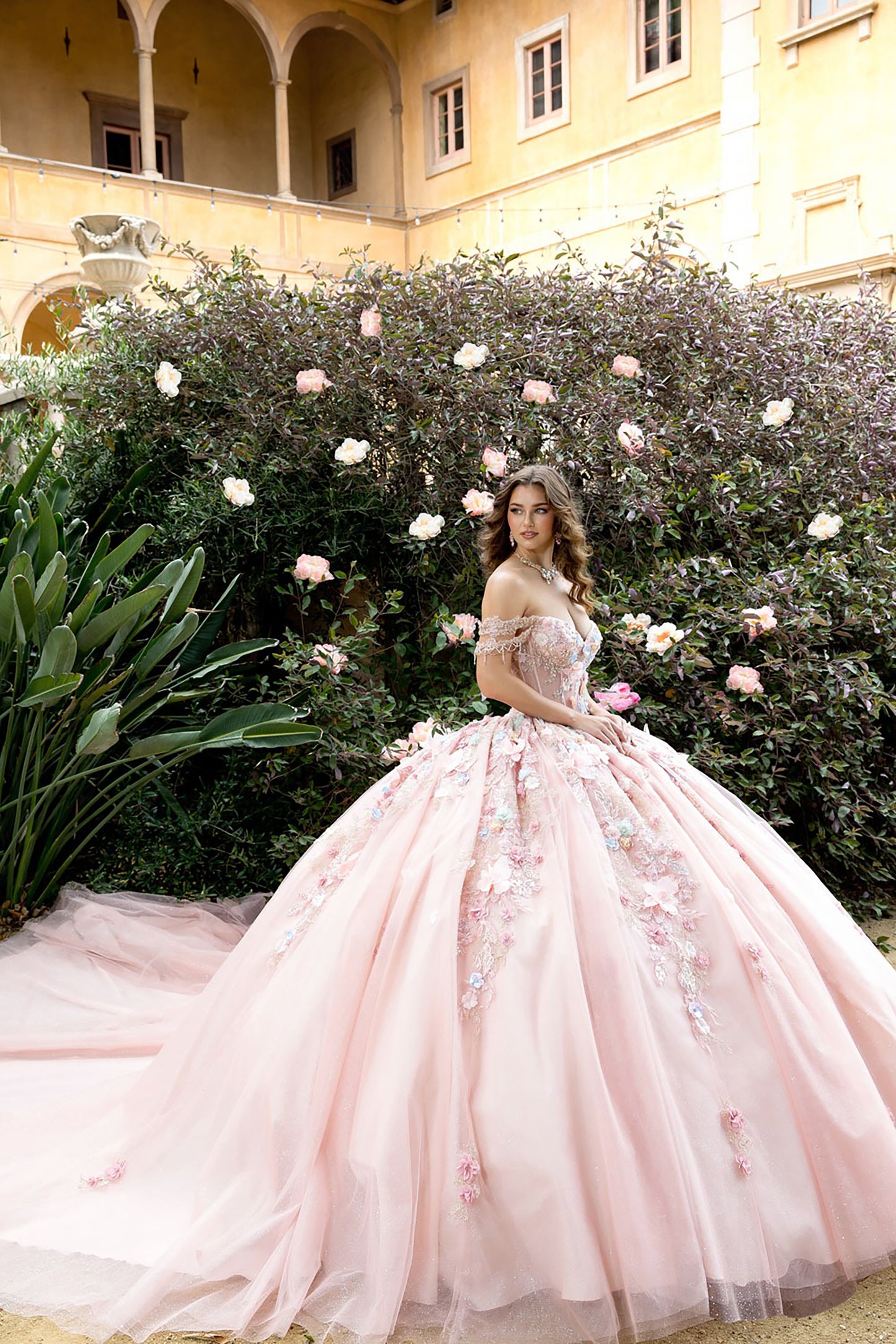 Woman in a pink floral gown standing in a garden with flowers and a building in the background