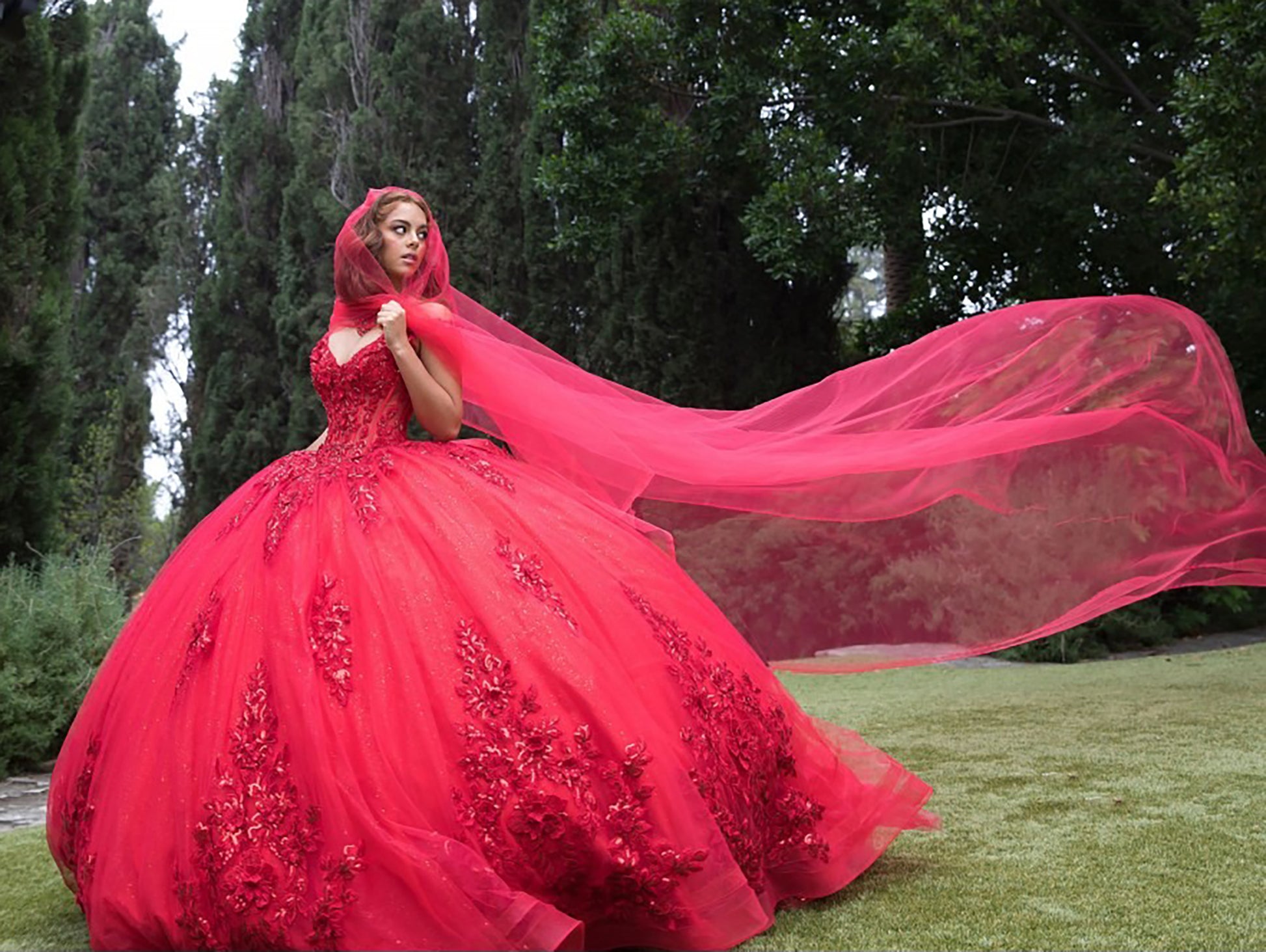 Woman in a red gown with a long train standing outdoors with trees in the background