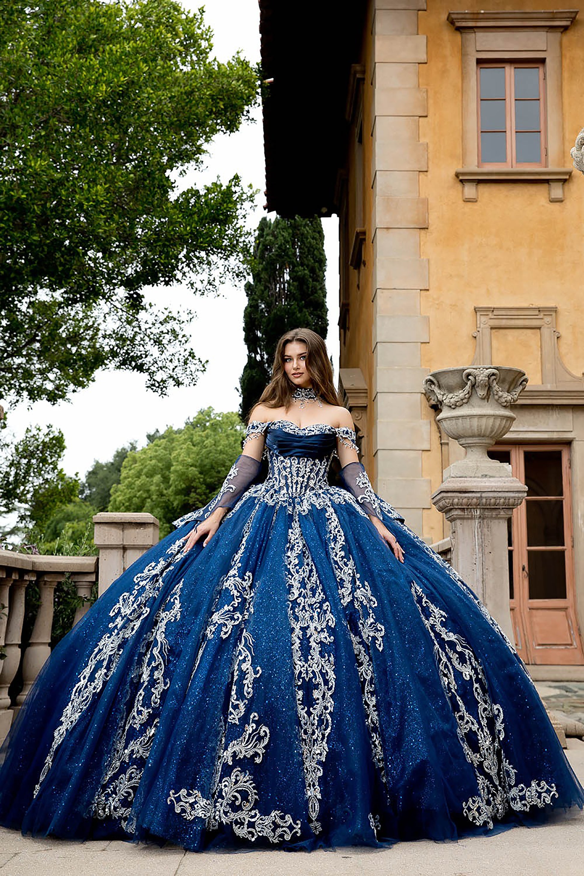 Woman in a blue ball gown with white embroidery standing in front of a building.
