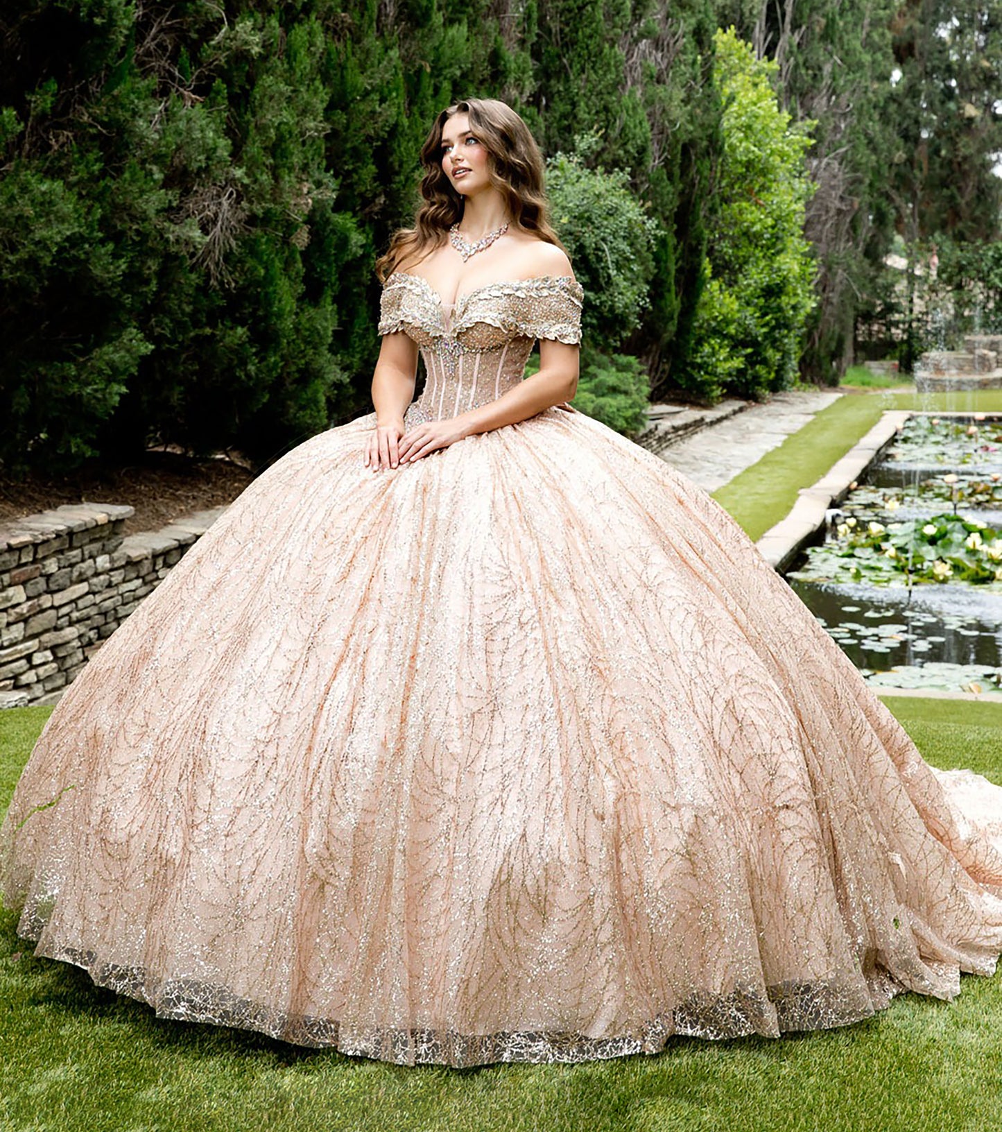 Woman in a rose gold ball gown standing in a garden with greenery and water features.