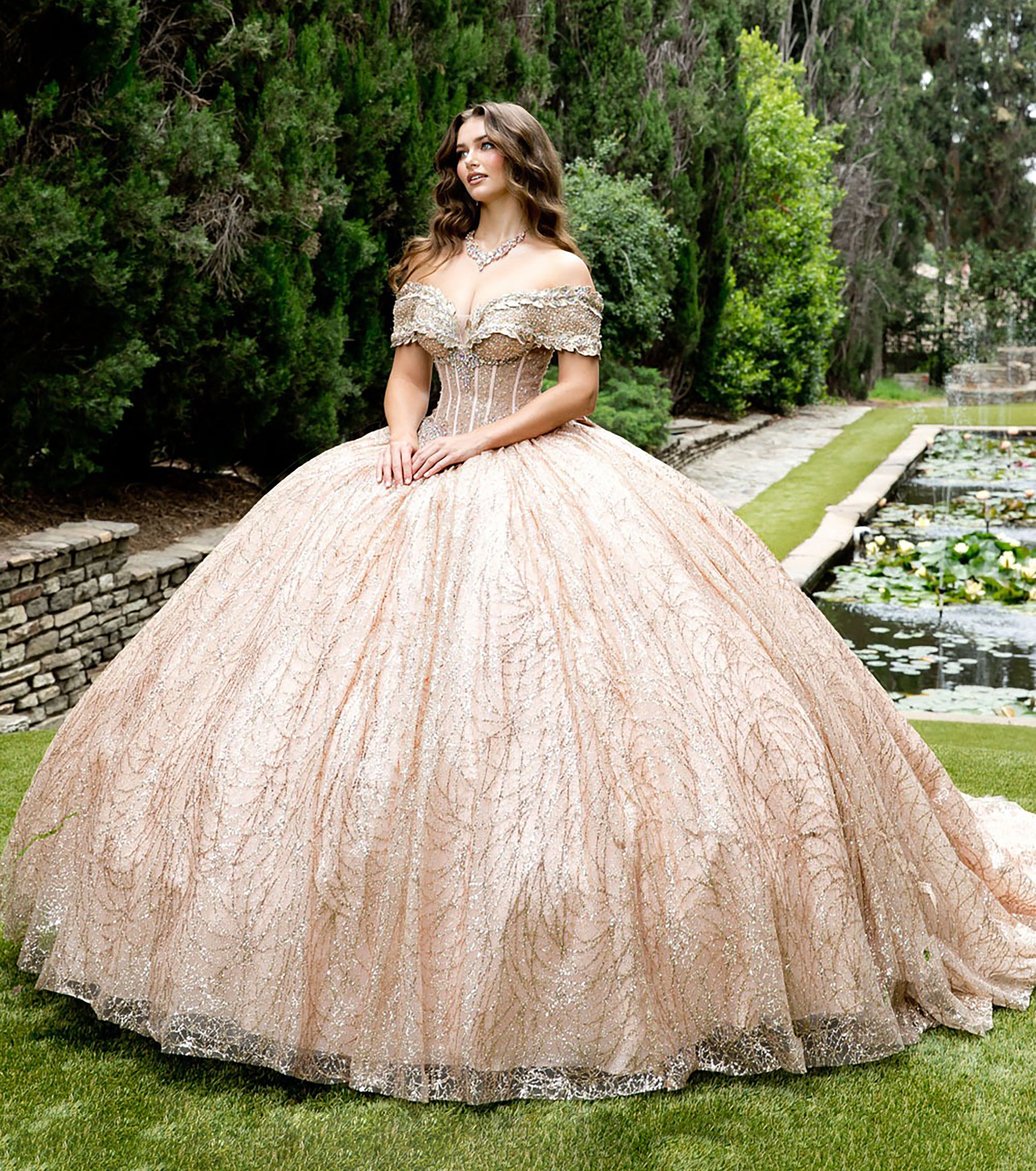 Woman in a rose gold ball gown standing in a garden with greenery and water features.