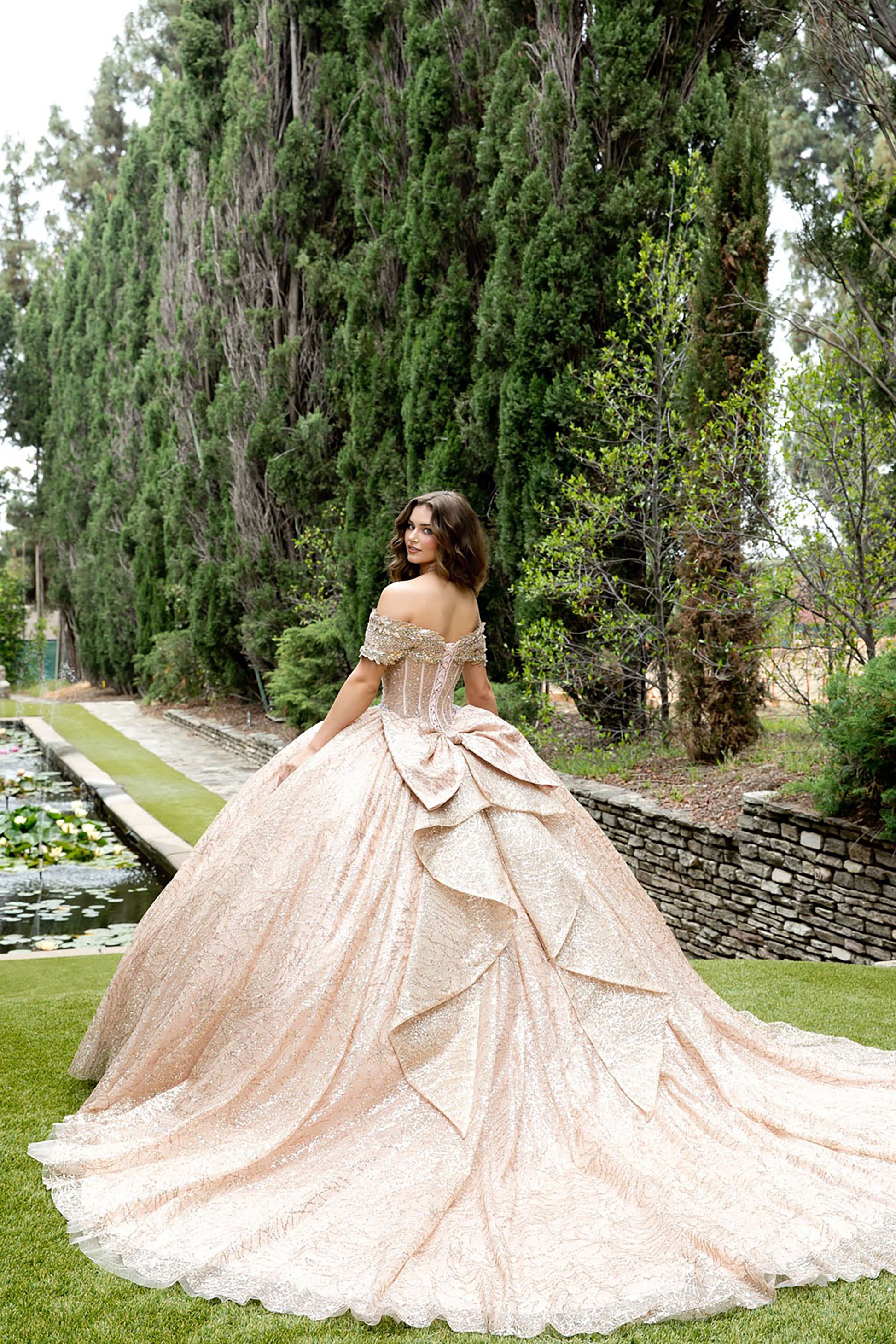 Woman in a glamorous off-shoulder gown standing in a garden with tall trees and stone walls.