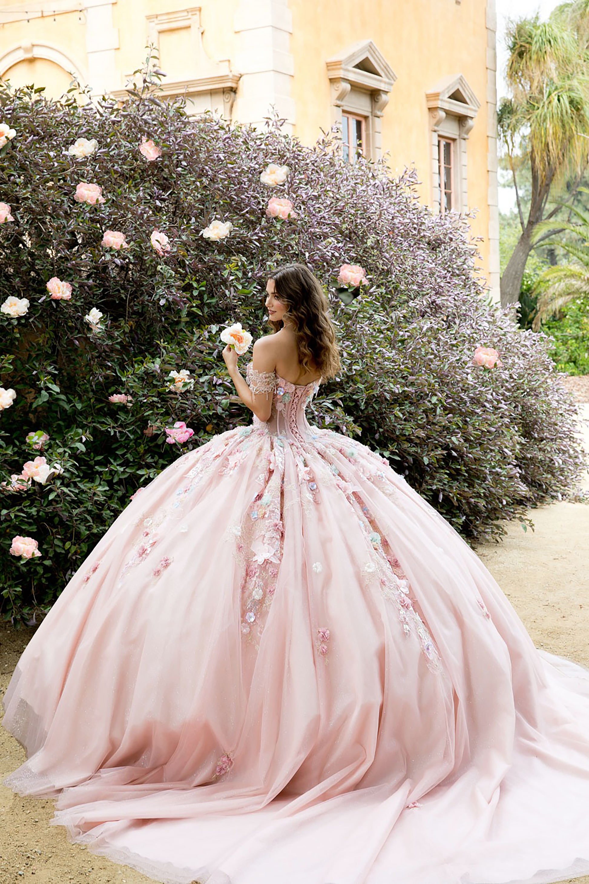 Woman in a pink gown standing in front of a floral bush with a building in the background