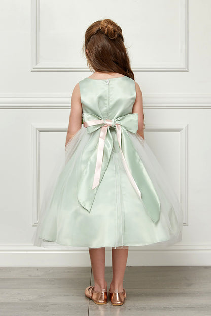 Young girl wearing a light green dress with a bow standing against a white paneled wall.