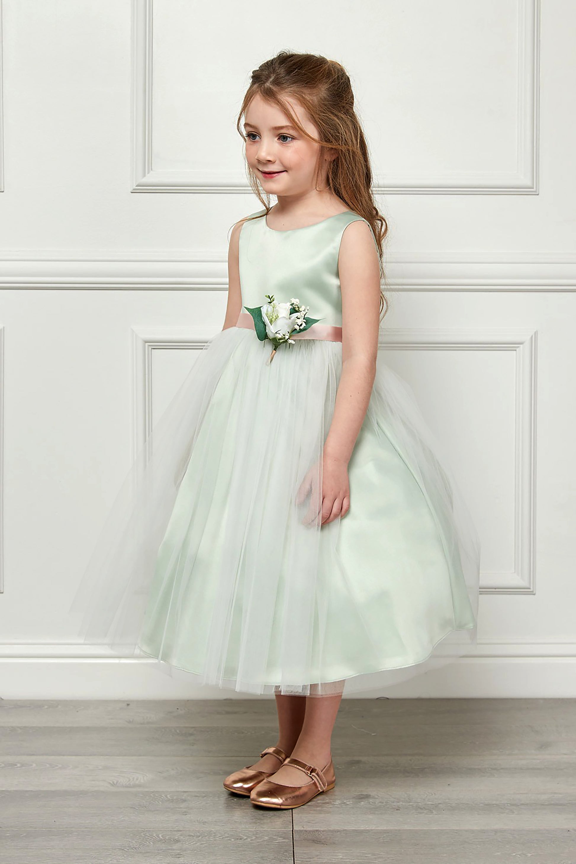 Young girl in a light green dress with a floral corsage standing against a white paneled wall.