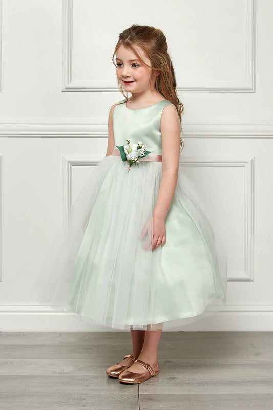 Young girl in a light green dress with a floral corsage standing against a white paneled wall.