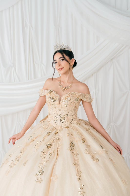 Woman in a champagne off-shoulder gown with floral details against a white textured wall.