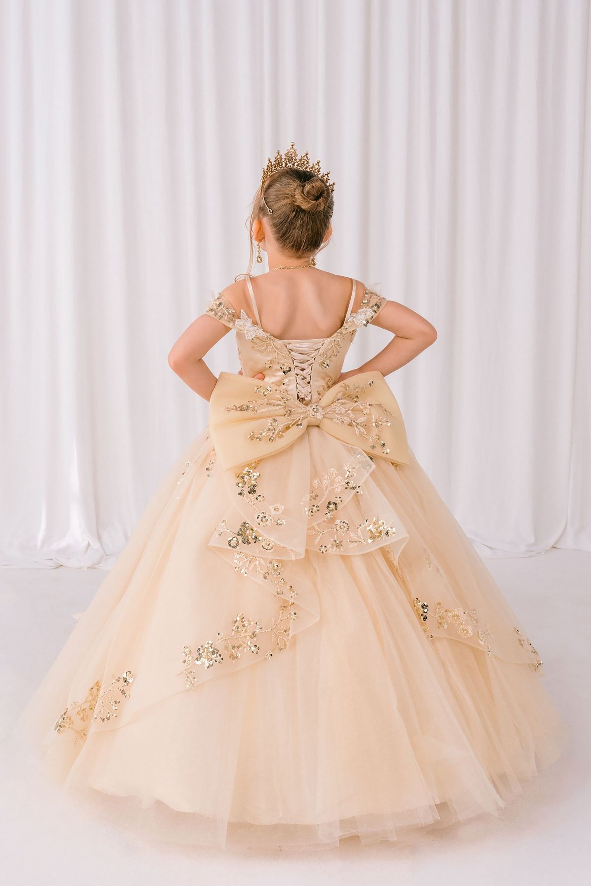 Young girl in a beige ball gown with floral embroidery and a large bow at the waist, standing against a white curtain.