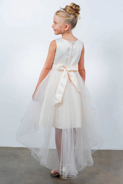 Young girl standing back-facing in champagne sleeveless high-low tulle flower girl dress with lace-up closure, pearl trim neckline, rhinestone details, and cascading skirt