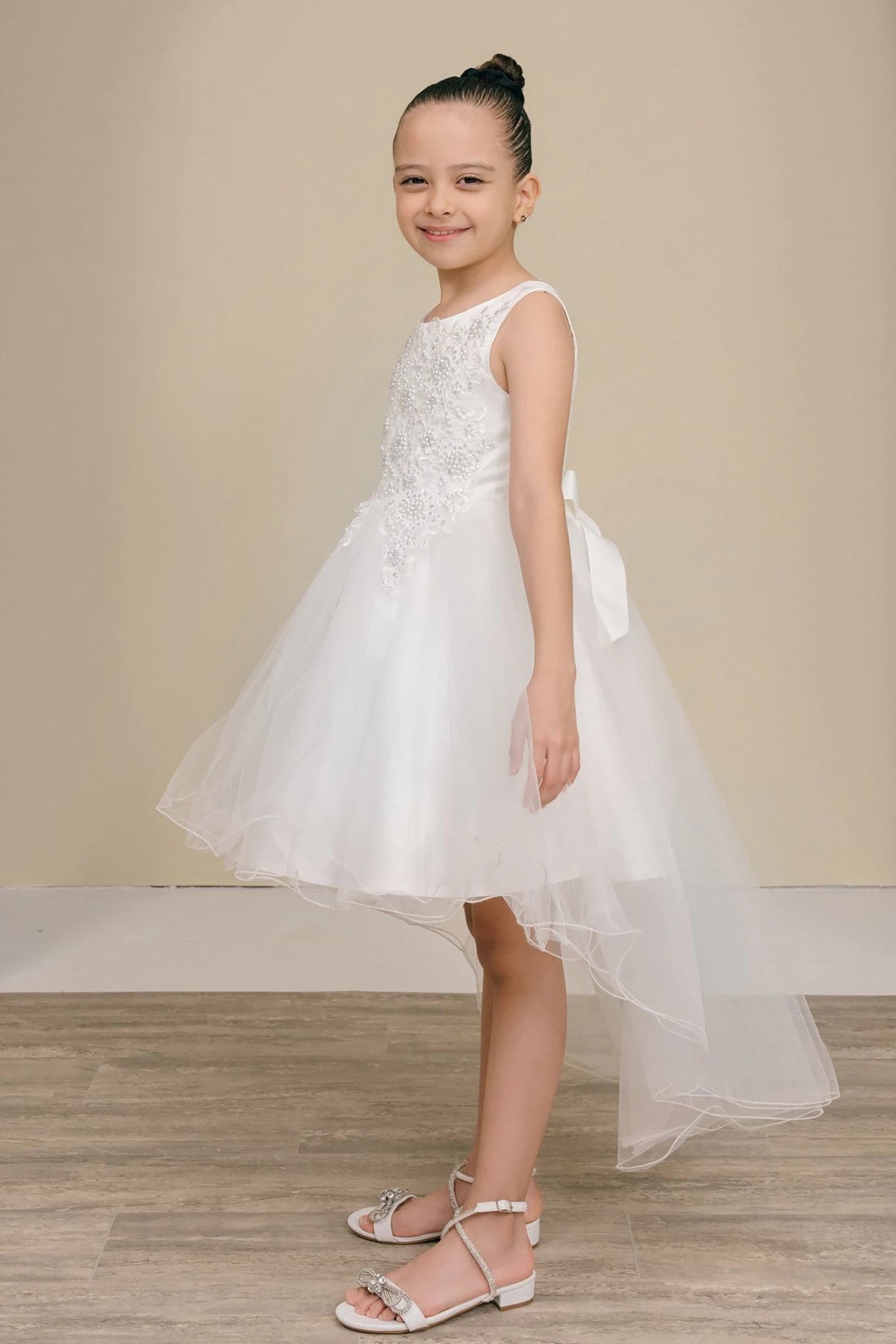 Young girl standing side-facing in white sleeveless high-low tulle flower girl dress with lace detailing, pearl accents, rhinestone sparkle, and flowing layered skirt