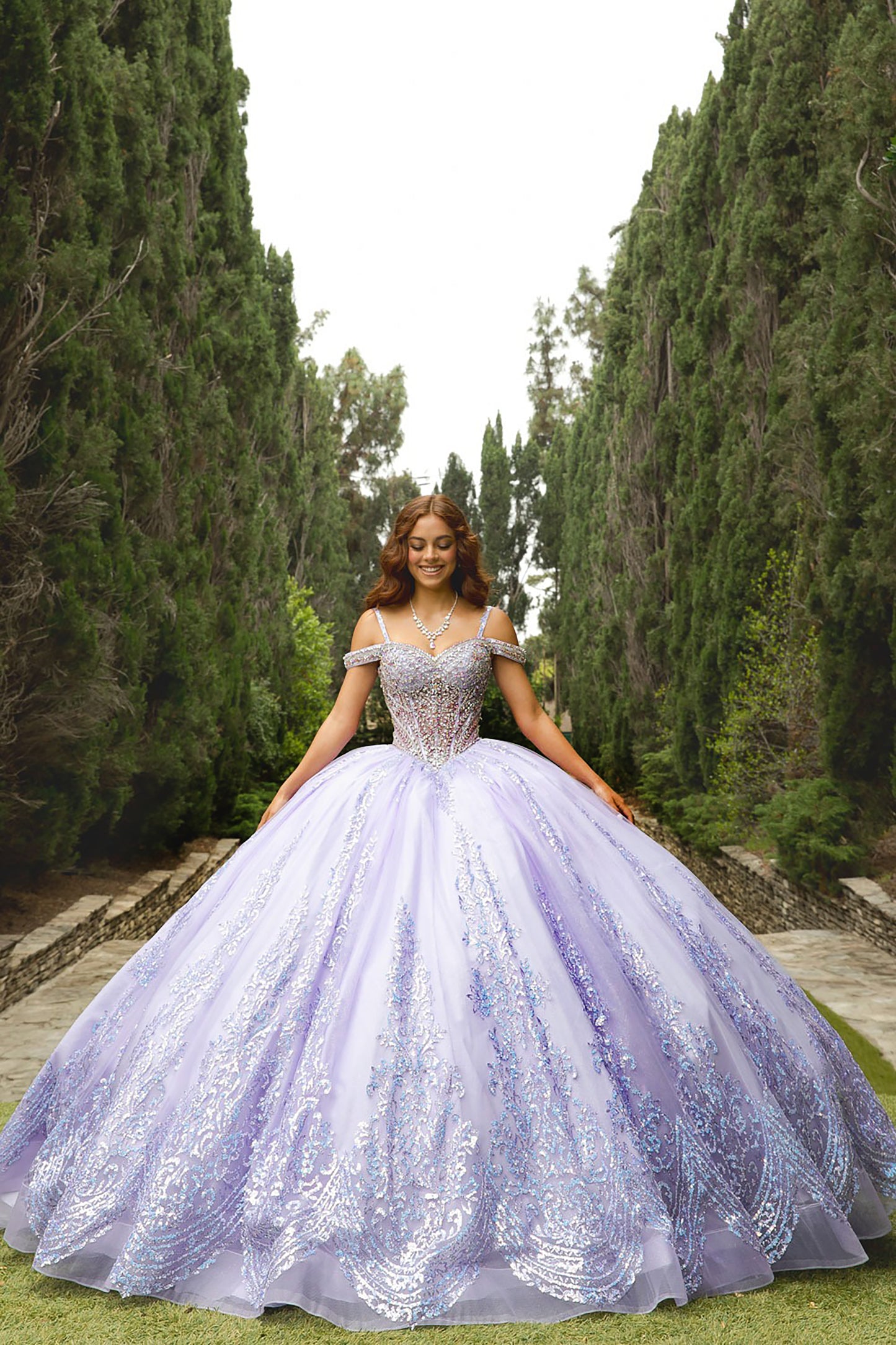 Woman in a purple and white floral dress standing in a garden with tall trees.