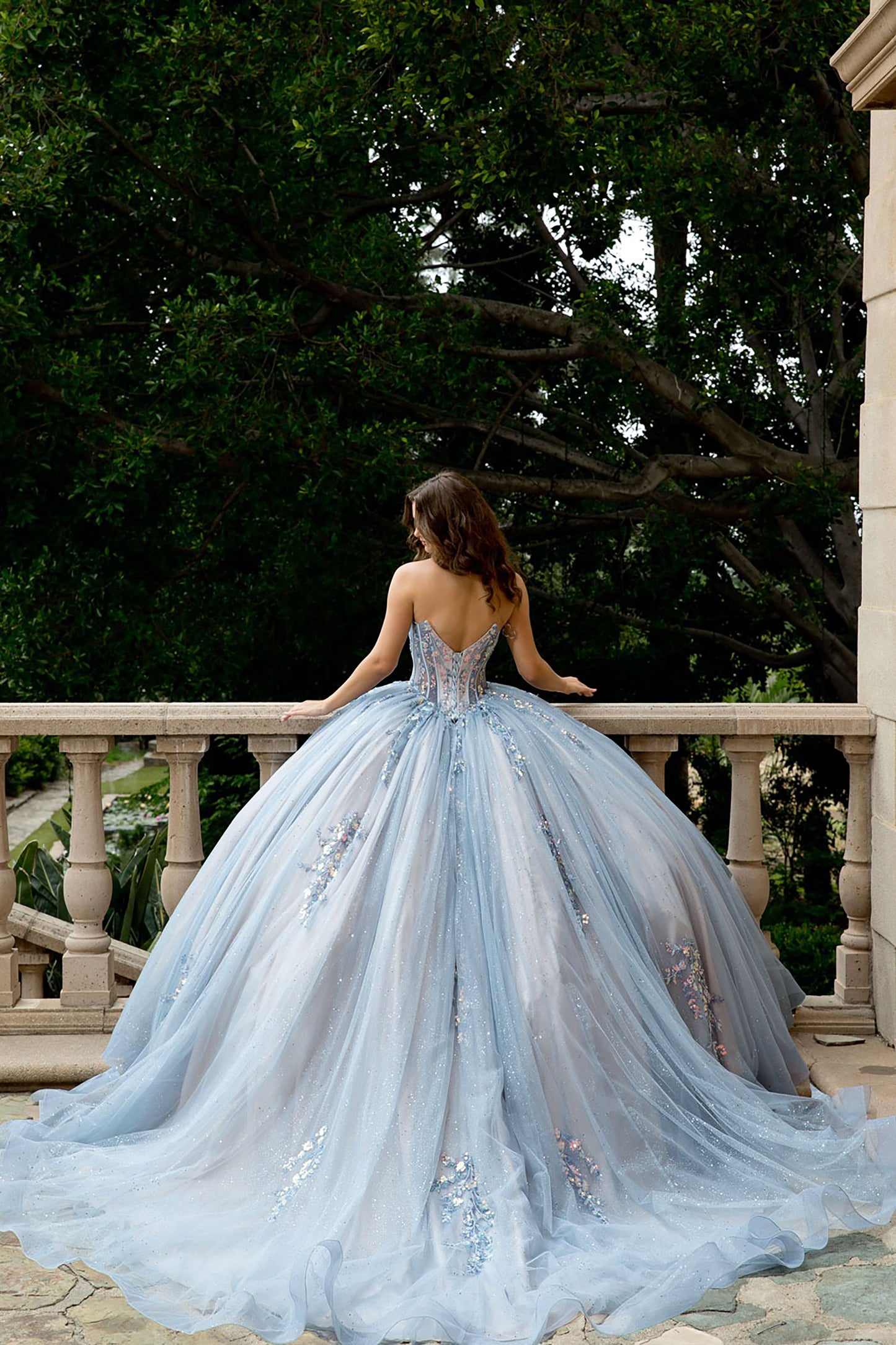Woman in a light blue gown standing on a balcony with trees in the background