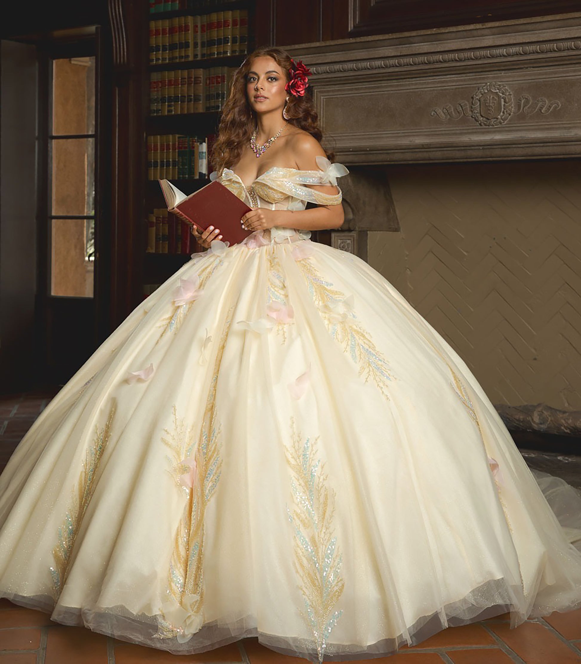 Woman in a white gown with floral details holding a red book in a room with a fireplace.