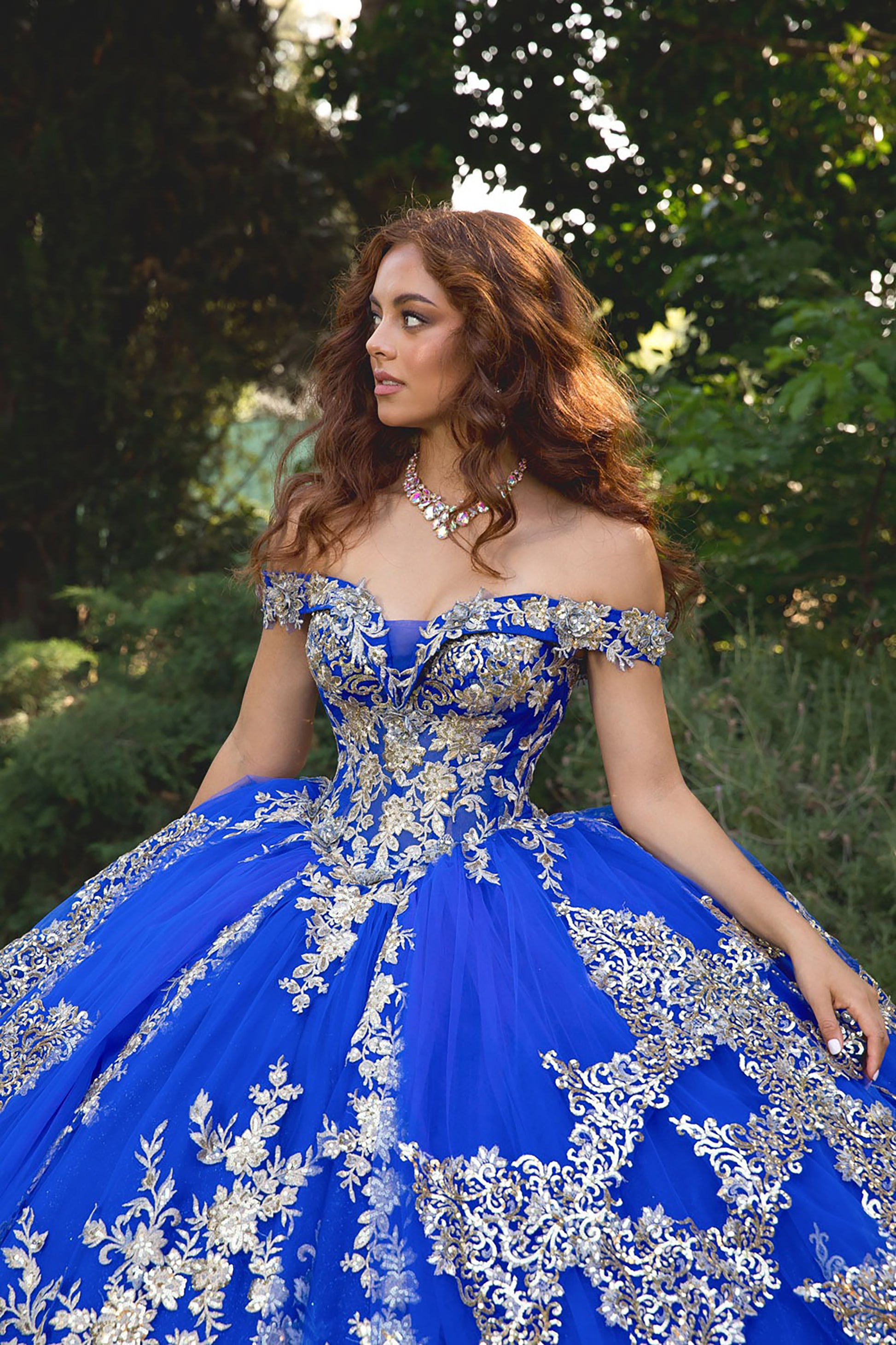 Woman wearing a blue and silver formal gown with floral embroidery against a natural background.