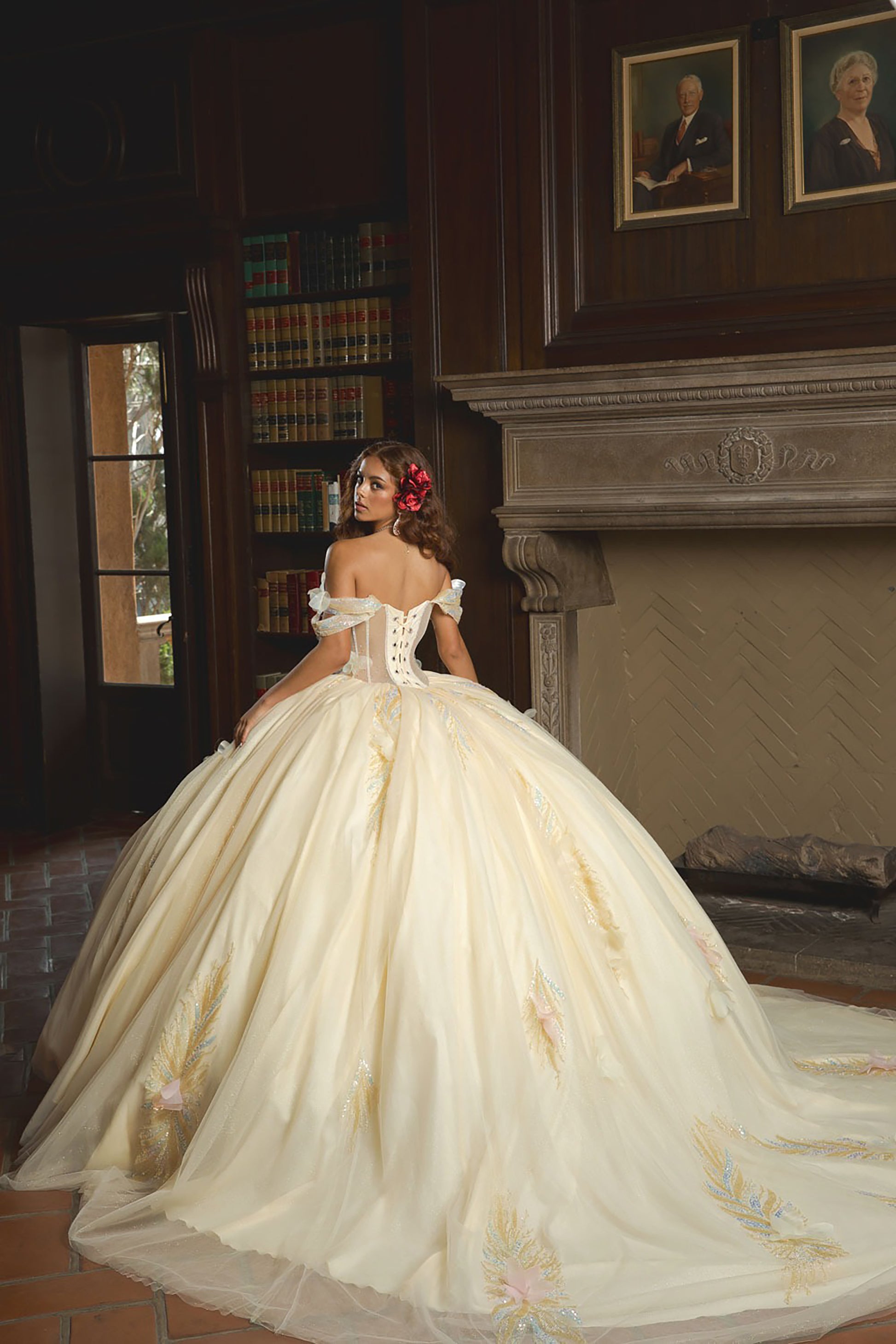 Woman in a white wedding dress with floral details in a room with bookshelves and portraits.