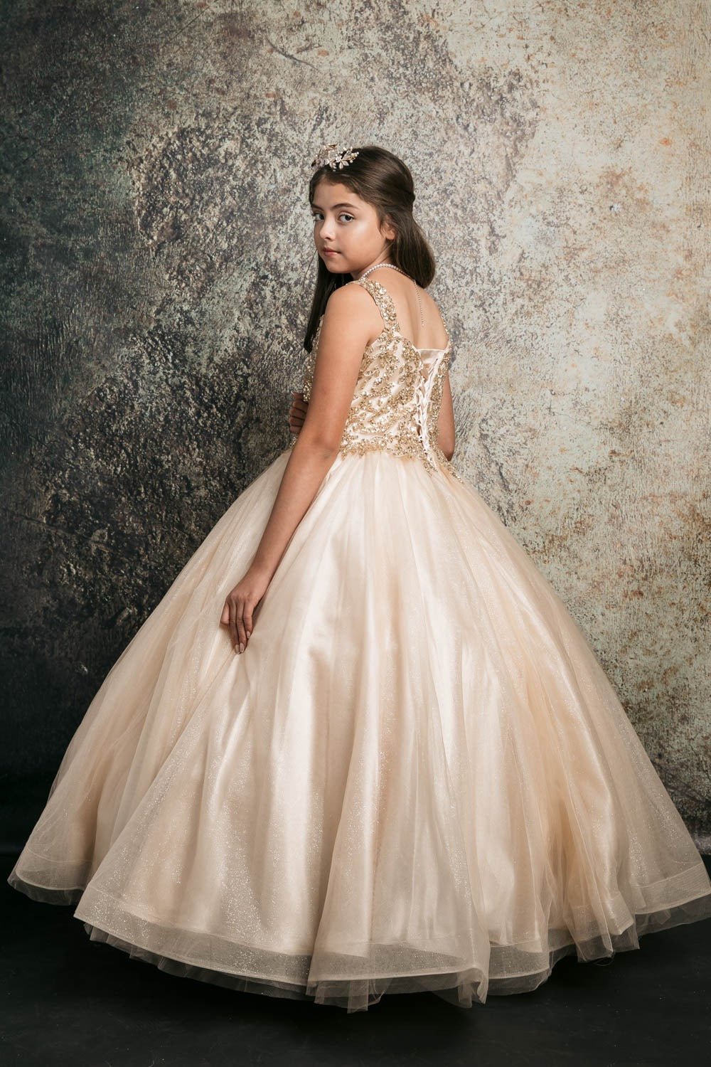 Young girl standing back-facing in champagne kids mini quinceañera dress with sequin beaded embroidery bodice and glitter tulle layered skirt